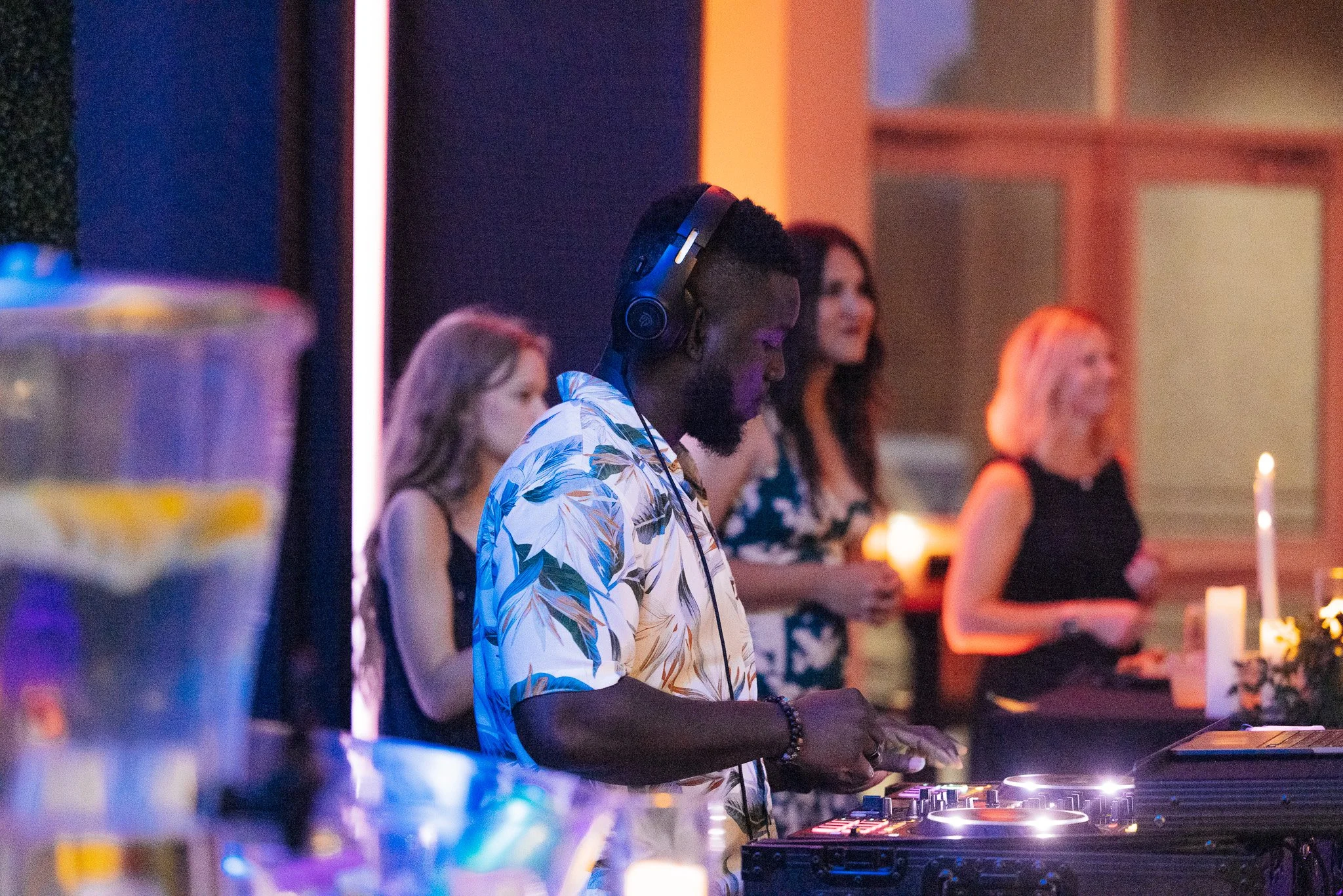 A DJ with a beard and wearing a floral shirt, headphones, and a beaded bracelet, is DJing at a party with women in the background, some holding drinks, in a dimly lit indoor setting.
