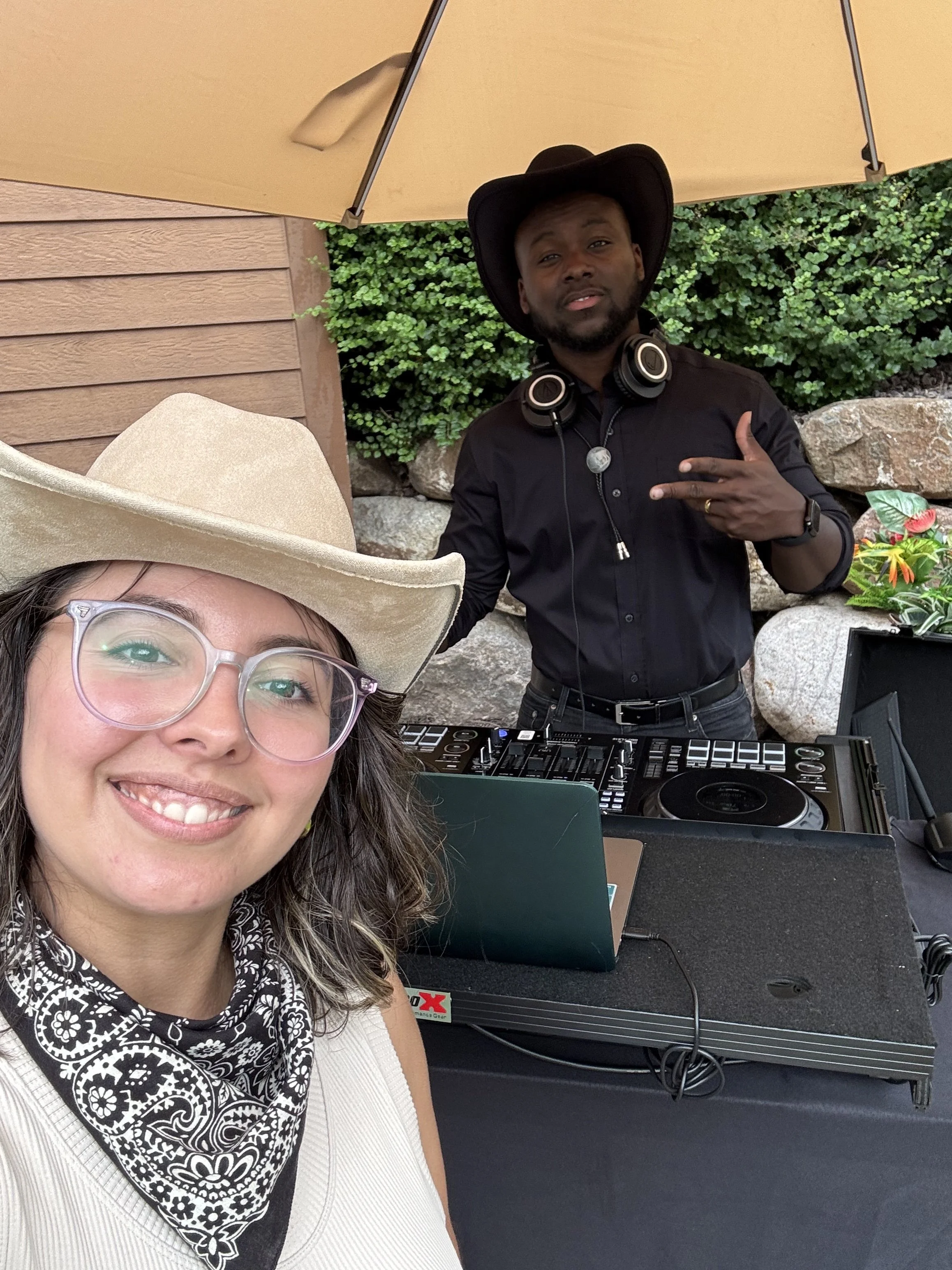 A woman with glasses and a beige cowboy hat smiling at the camera, taking a selfie with a man in the background. The man is wearing a black hat, black shirt, and headphones around his neck, standing behind a DJ setup with a laptop and equipment, unde
