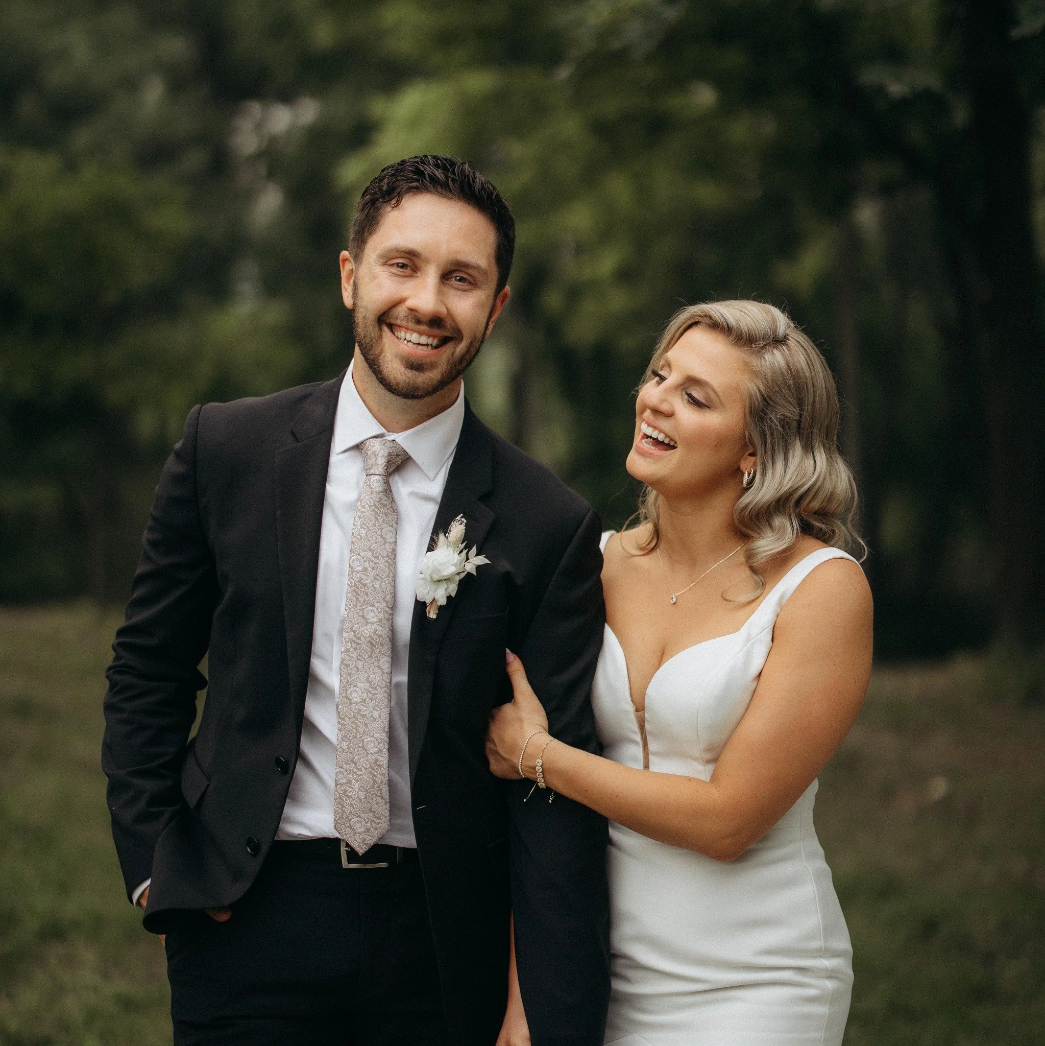 A smiling man in a black suit and patterned tie standing outdoors with a woman in a white dress, holding the man’s arm, both appearing happy and dressed for a wedding or special occasion, with trees and greenery in the background.
