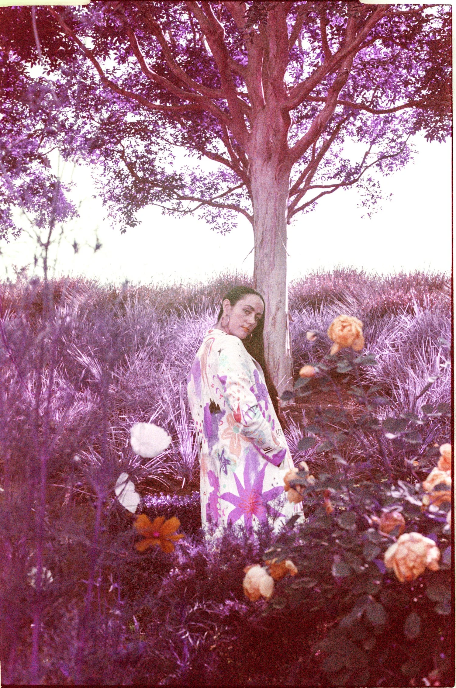 A woman with long dark hair, wearing a floral jacket, standing in a rose garden with a large tree visible in the background. The image has a pinkish hue.