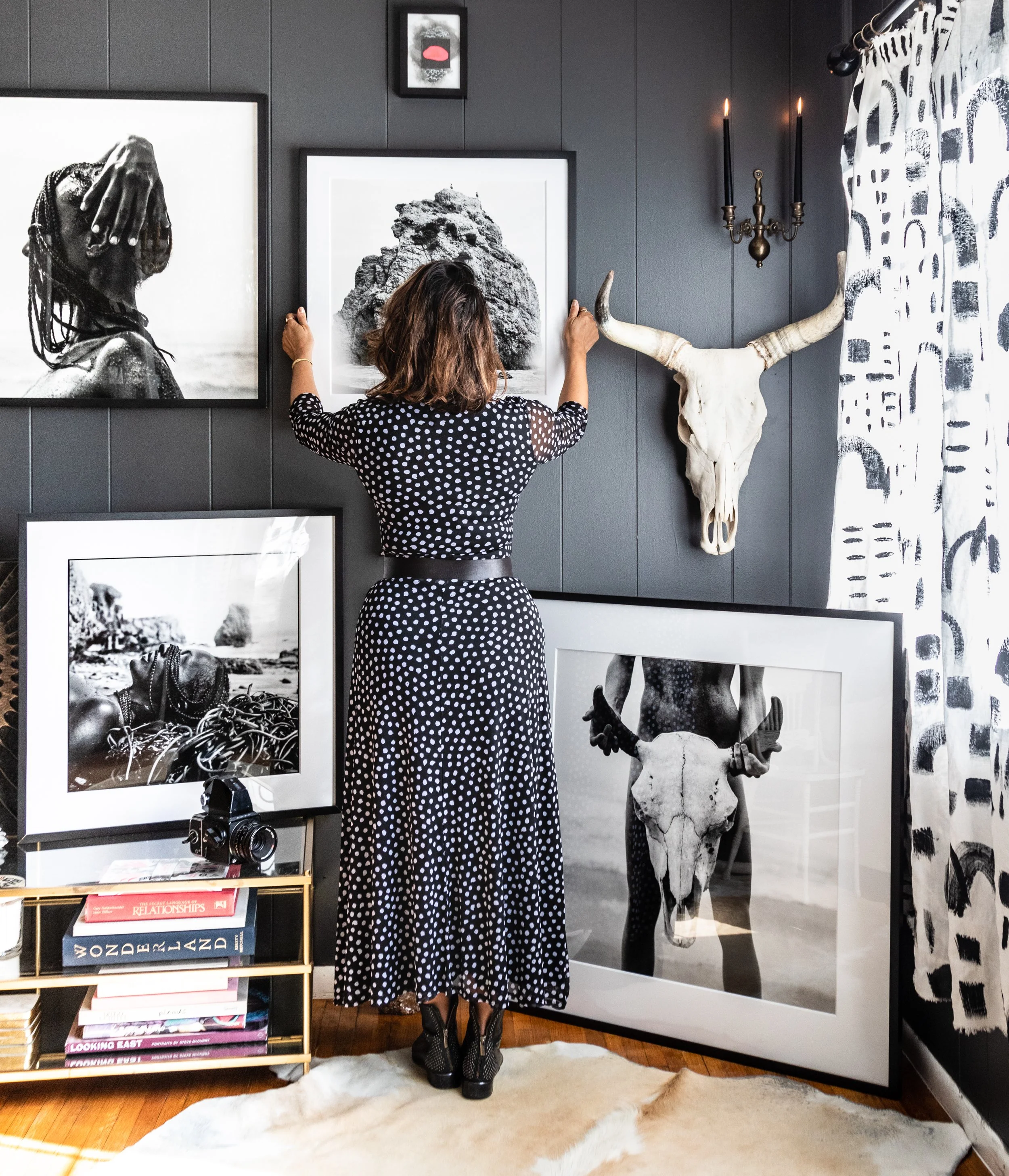Woman in a black and white polka dot dress arranging black and white framed photographs on a dark wall in a stylish room with eclectic decor.