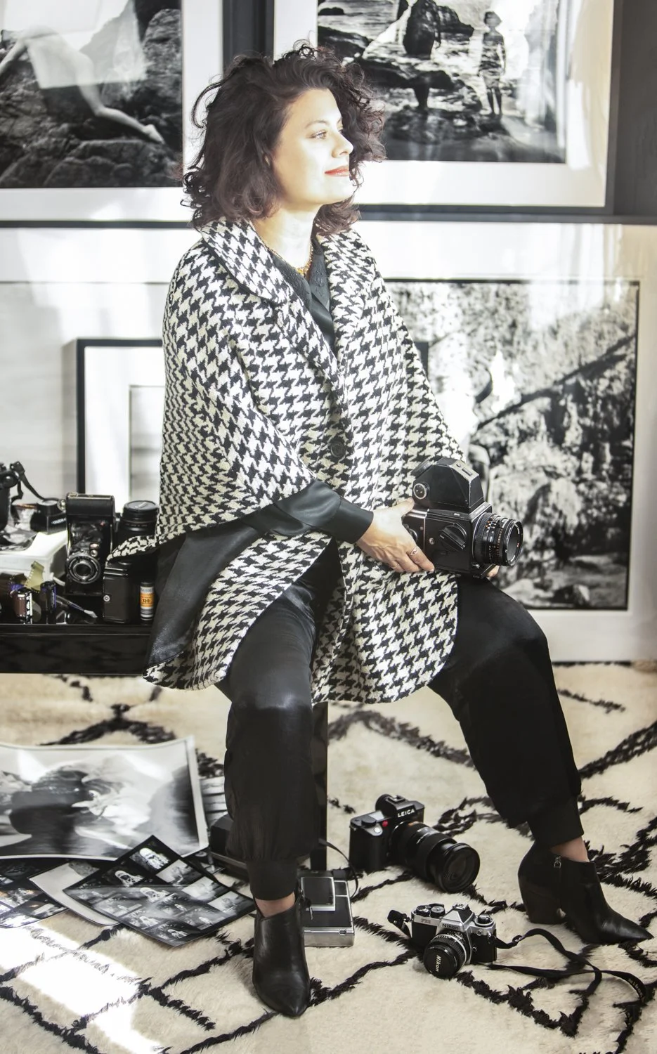 A woman sitting on a bench holding a vintage camera in a gallery with black and white photographs on the wall behind her. There are multiple cameras and photographs on the floor and a bench beside her.