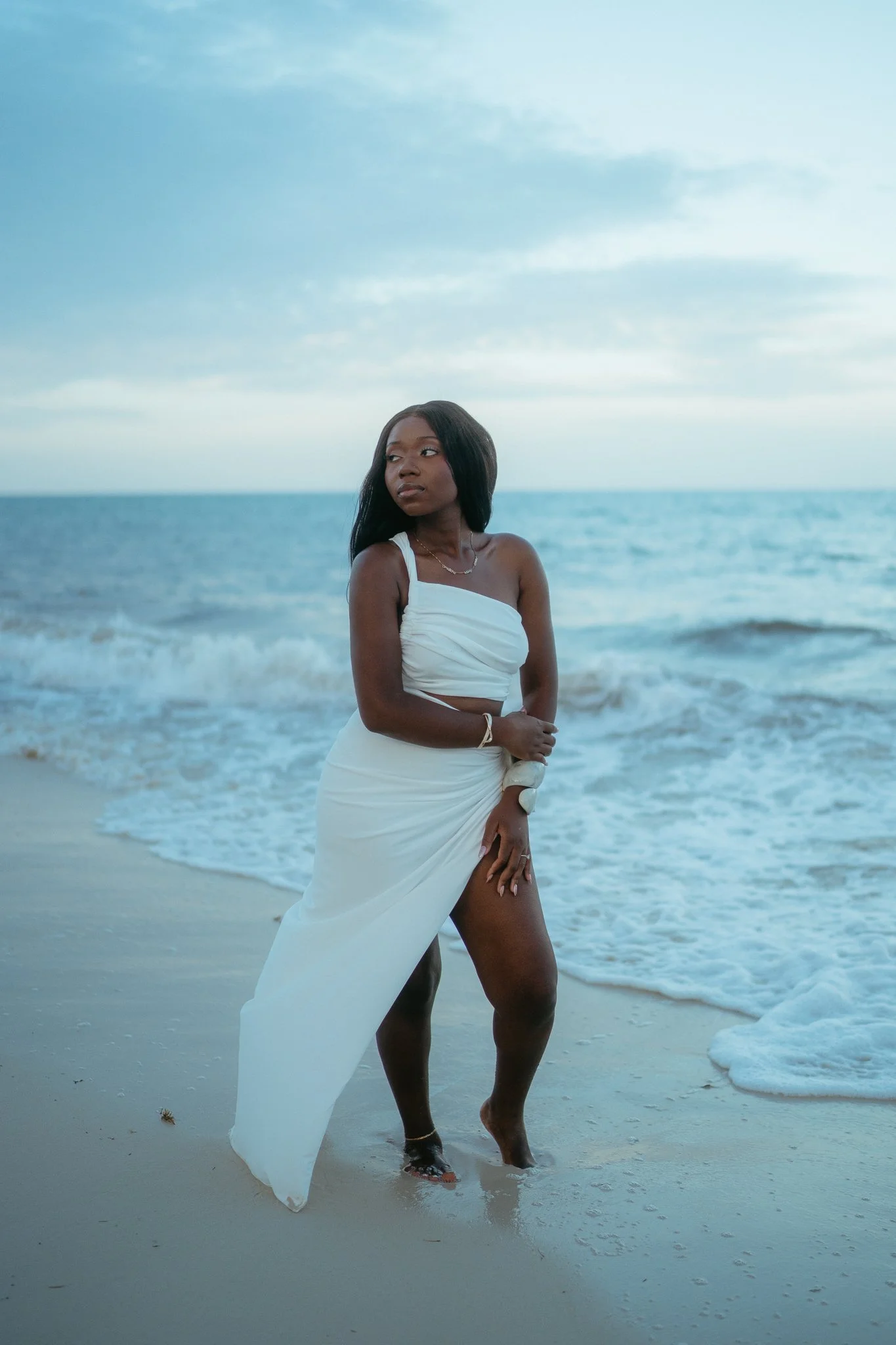 Mujer de piel oscura en la playa, vestida con un vestido blanco, con vista al mar y el cielo nublado.
