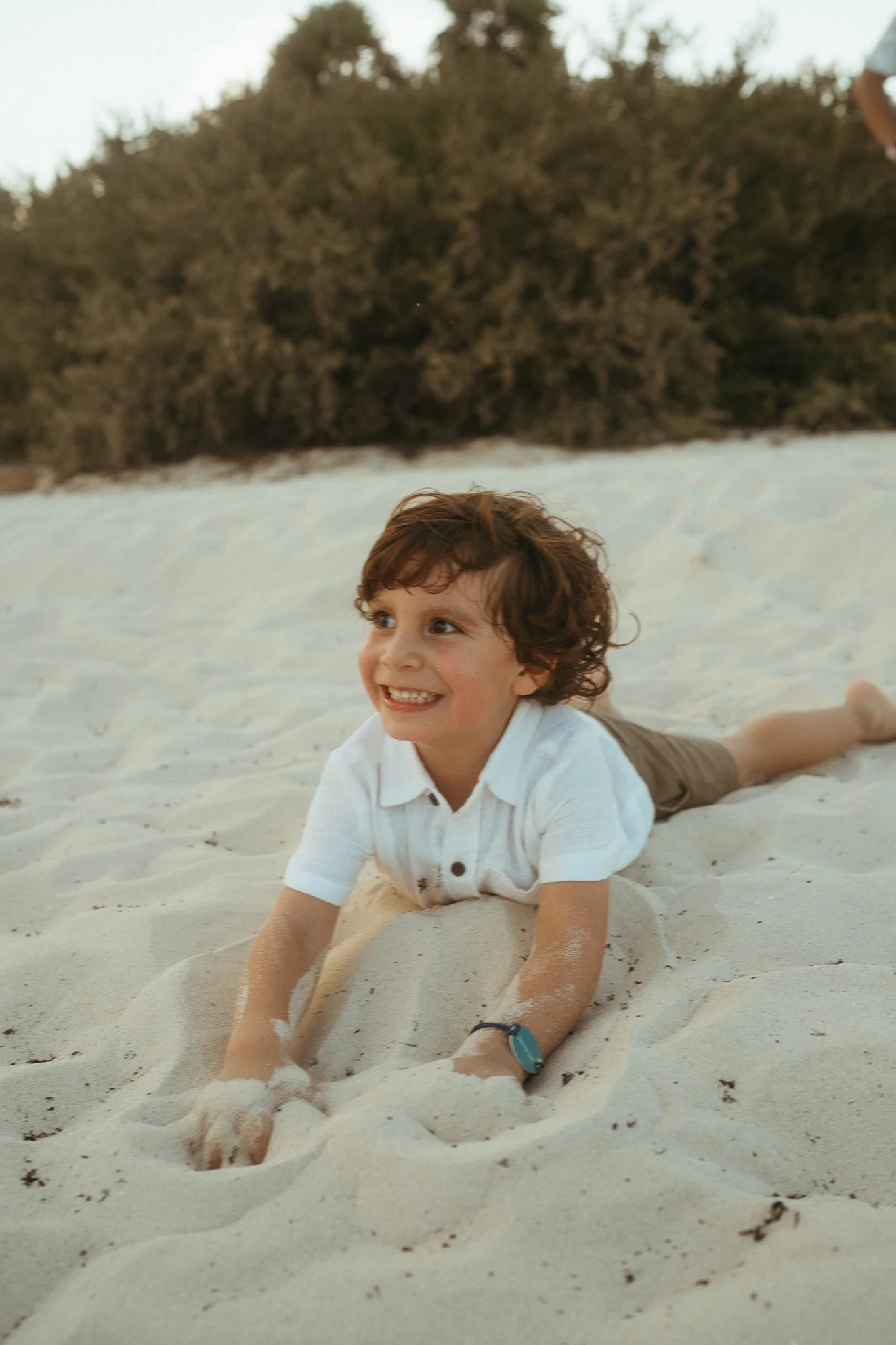 Niño sonriendo en la arena de la playa, con fondo de arbustos y cielo