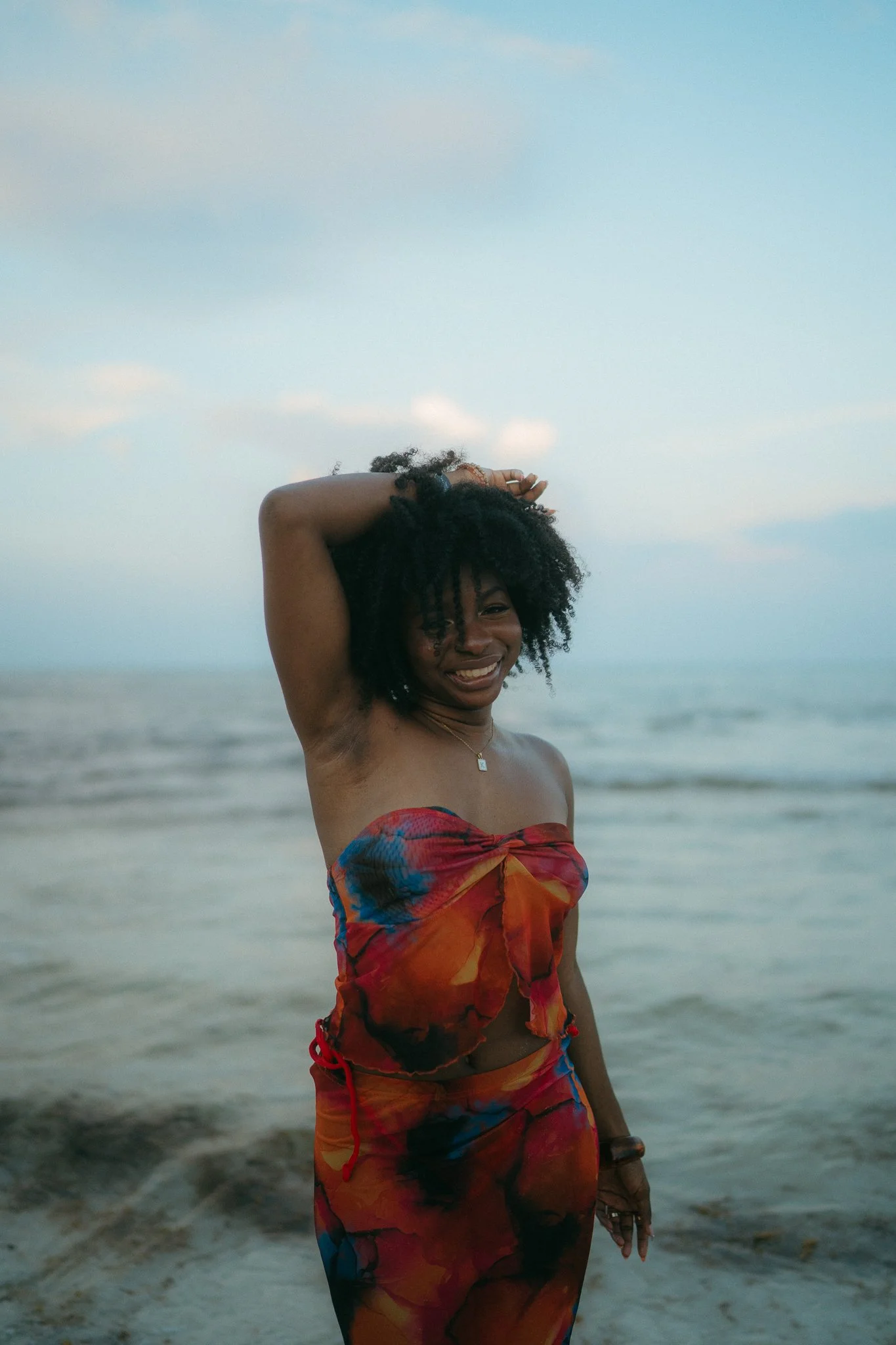 Mujer sonriendo en la playa con vestido colorido y cabello rizado, con el mar y cielo de fondo.