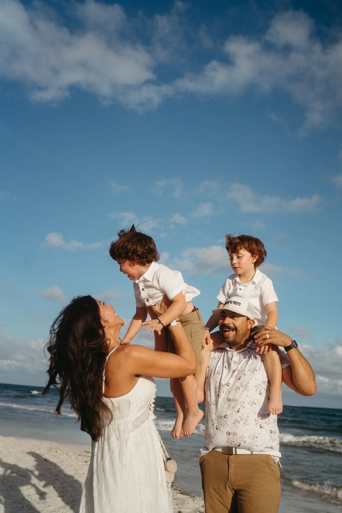 Familia en la playa disfrutando, dos niños en los hombros de los padres, risas y alegría.