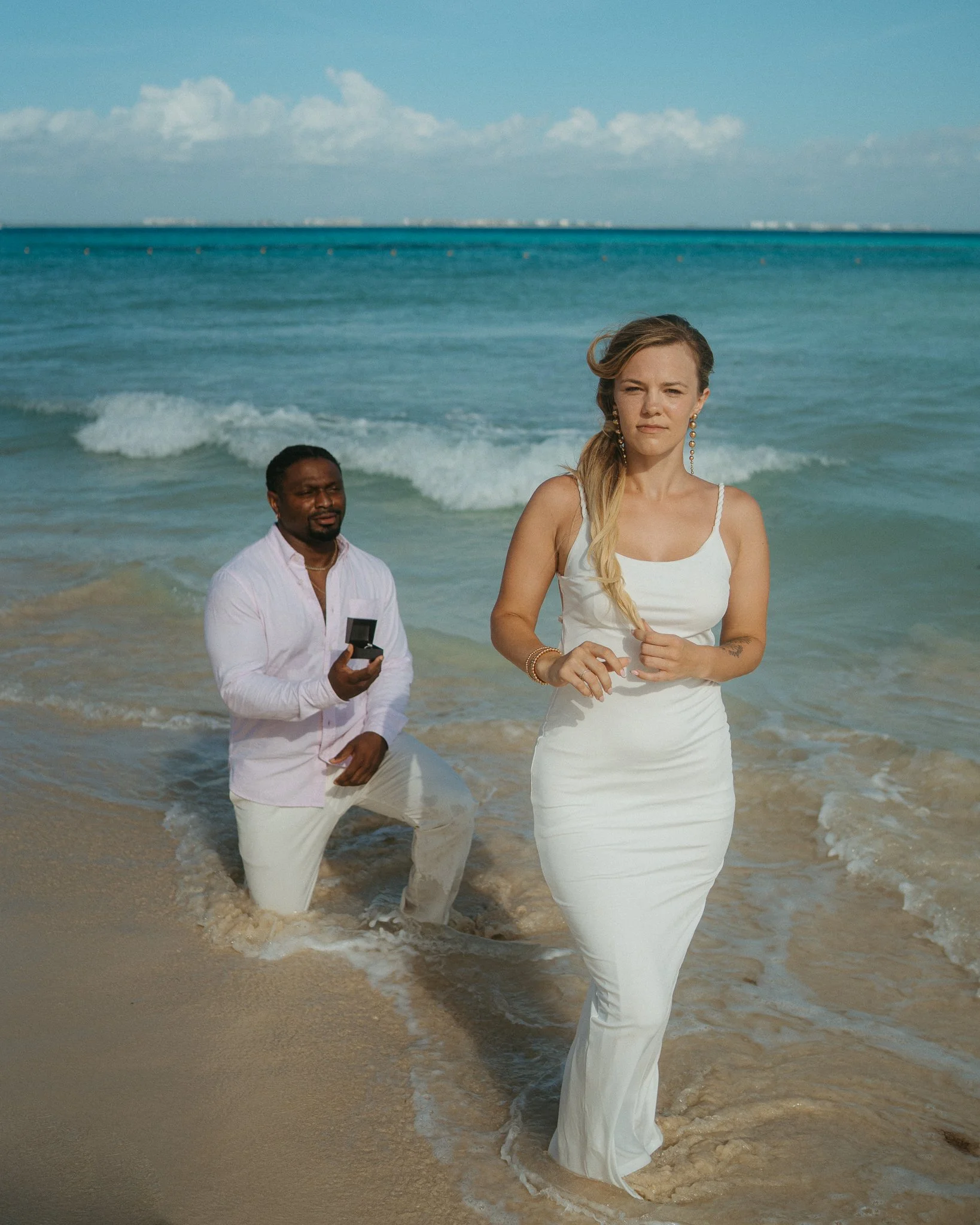 Una pareja en la playa, con el hombre arrodillado en la arena con una caja de anillo y la mujer de pie, en el agua, vestida de blanco, en una propuesta de matrimonio.