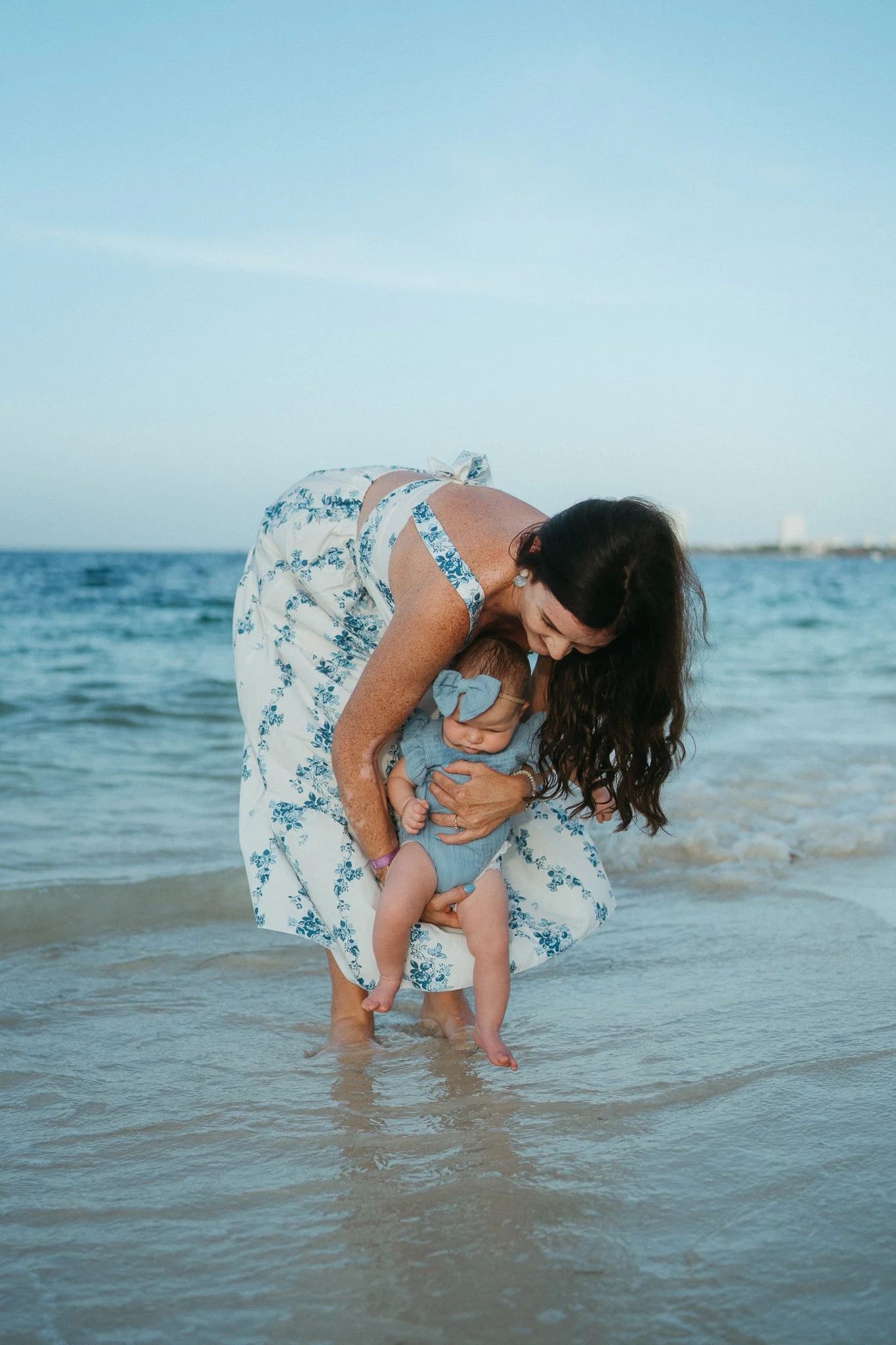 Mujer con vestido blanco y azul en la playa, sosteniendo a una bebé con un vestido azul y un moño en la cabeza, en el agua.