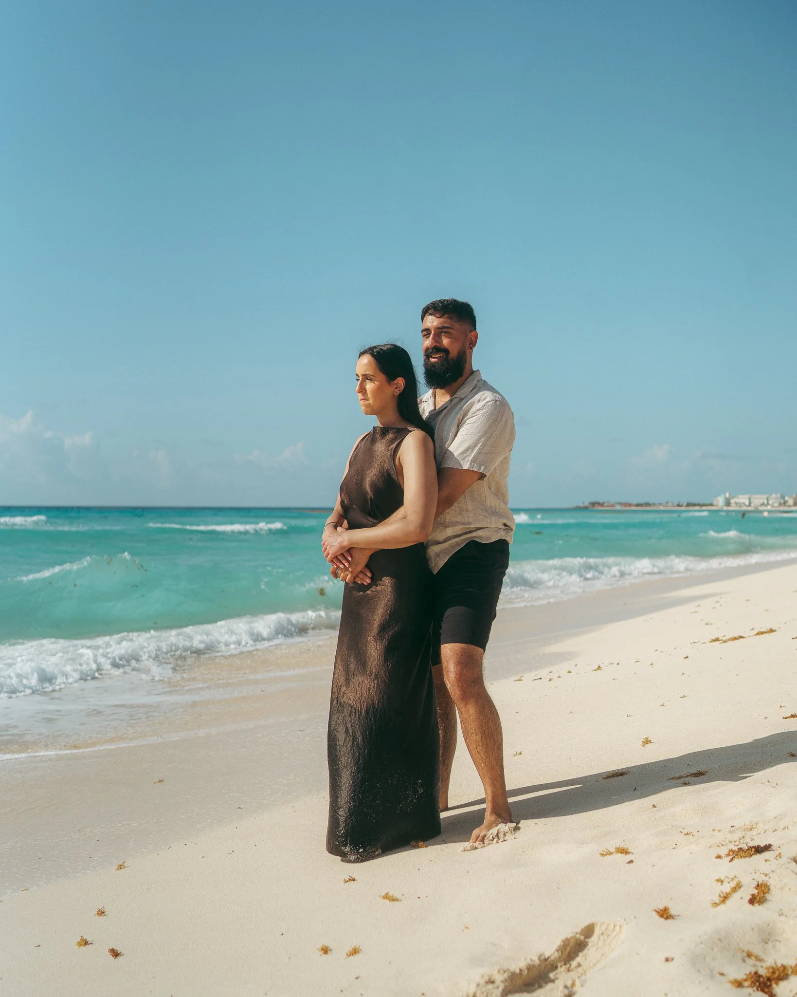 Una pareja en la playa, el hombre abraza a la mujer por detrás, ambos mirando al horizonte, con el mar y cielo despejado de fondo.