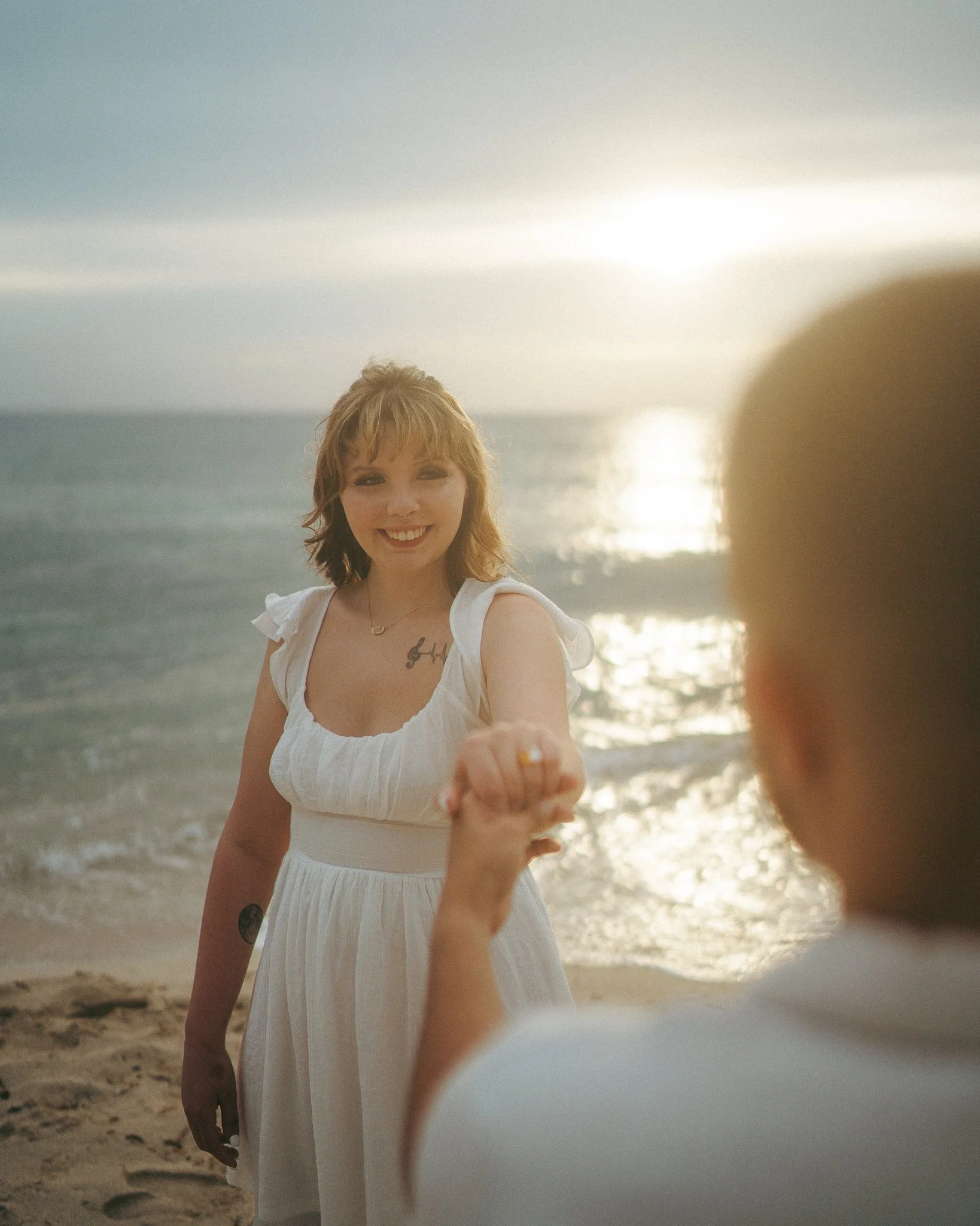 Una mujer con vestido blanco sonriendo en la playa, sosteniendo la mano a otra persona que no es visible en la imagen, durante un atardecer sobre el mar.