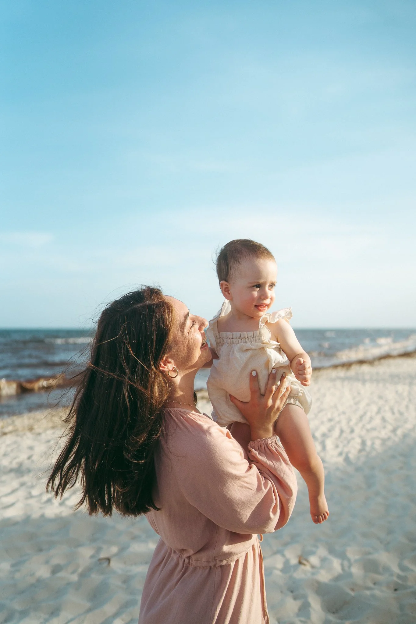 Una mujer con un vestido rosa sostiene en brazos a una niña en la playa, con el mar y el cielo azul de fondo.
