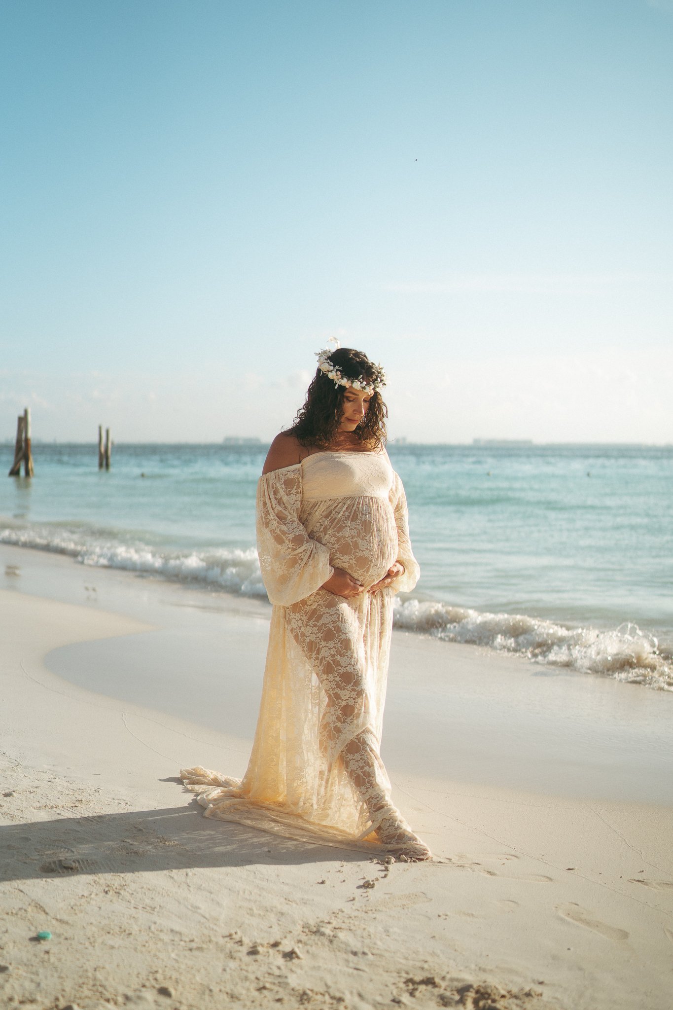 Mujer embarazada con vestido de encaje en la playa, sosteniendo su vientre, con corona de flores en la cabeza y mar en el fondo.