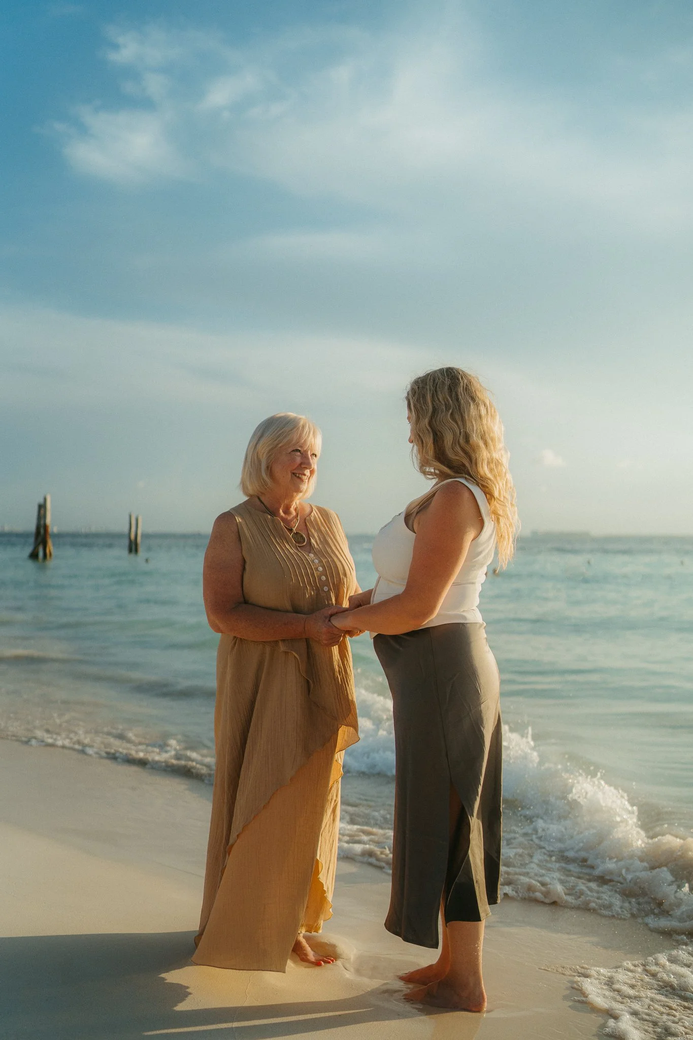Dos mujeres de diferentes edades sosteniéndose de las manos en la orilla del mar en una puesta de sol o atardecer, con olas suaves y cielo despejado.