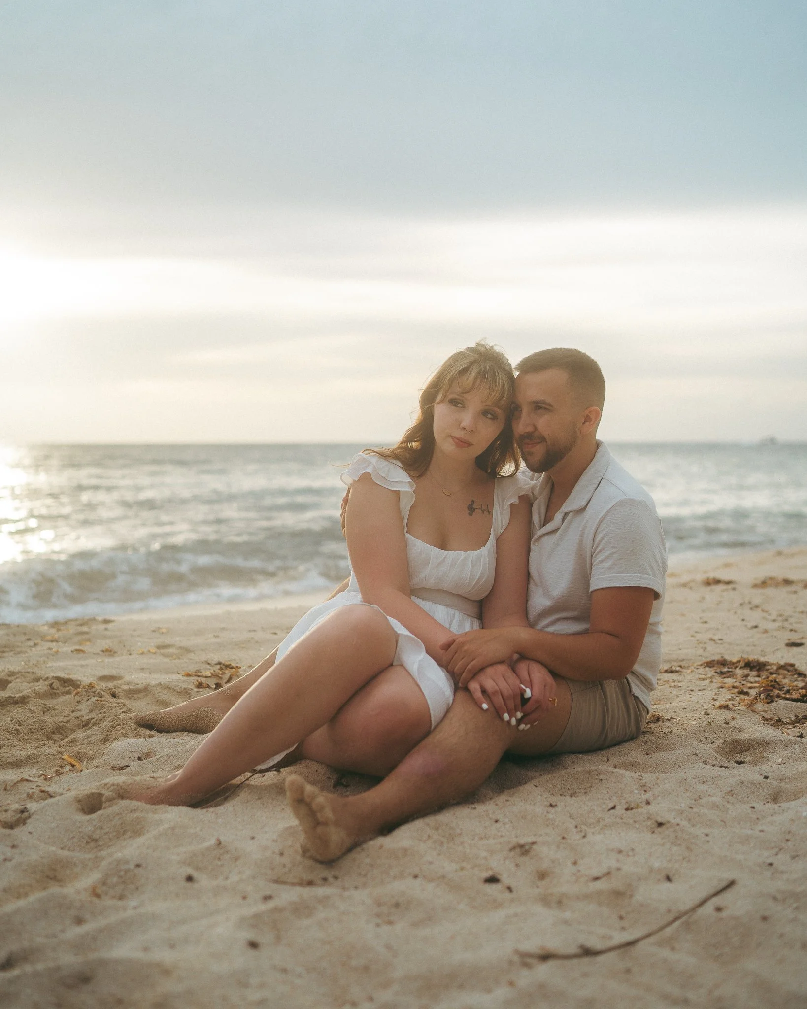 Una pareja sentada en la playa, con el mar y el cielo nublado al fondo, durante el atardecer.