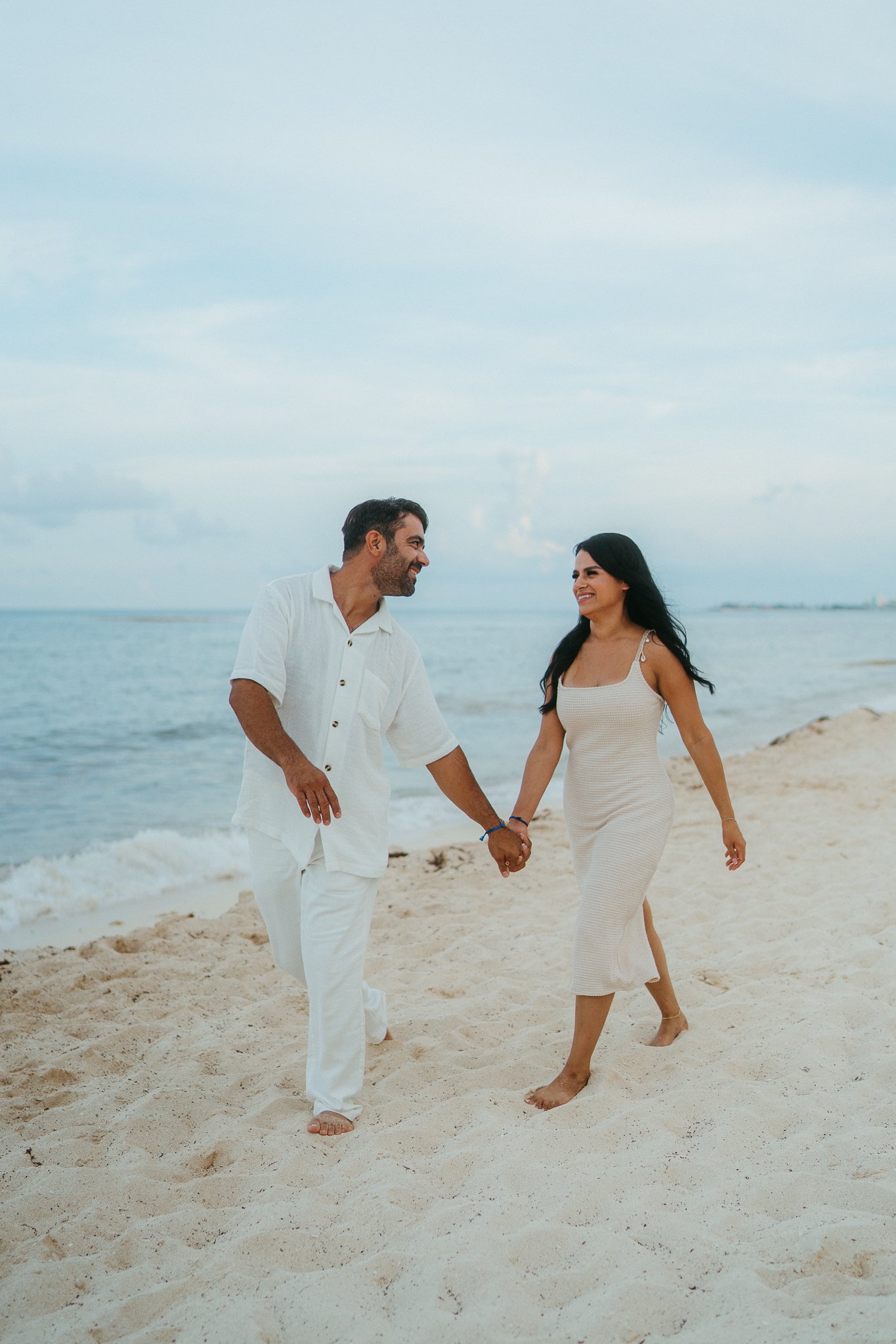 Una pareja caminando de la mano en la playa, riendo y disfrutando del momento, con el mar y el cielo en el fondo.