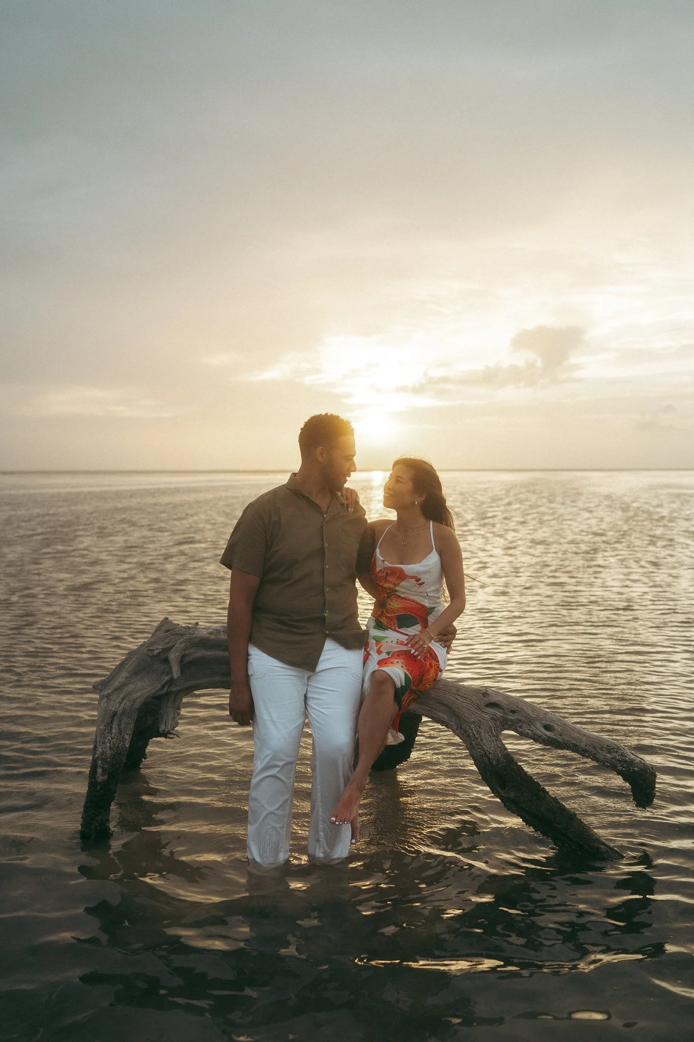 Una pareja en el agua al atardecer junto a un tronco caído en el mar.