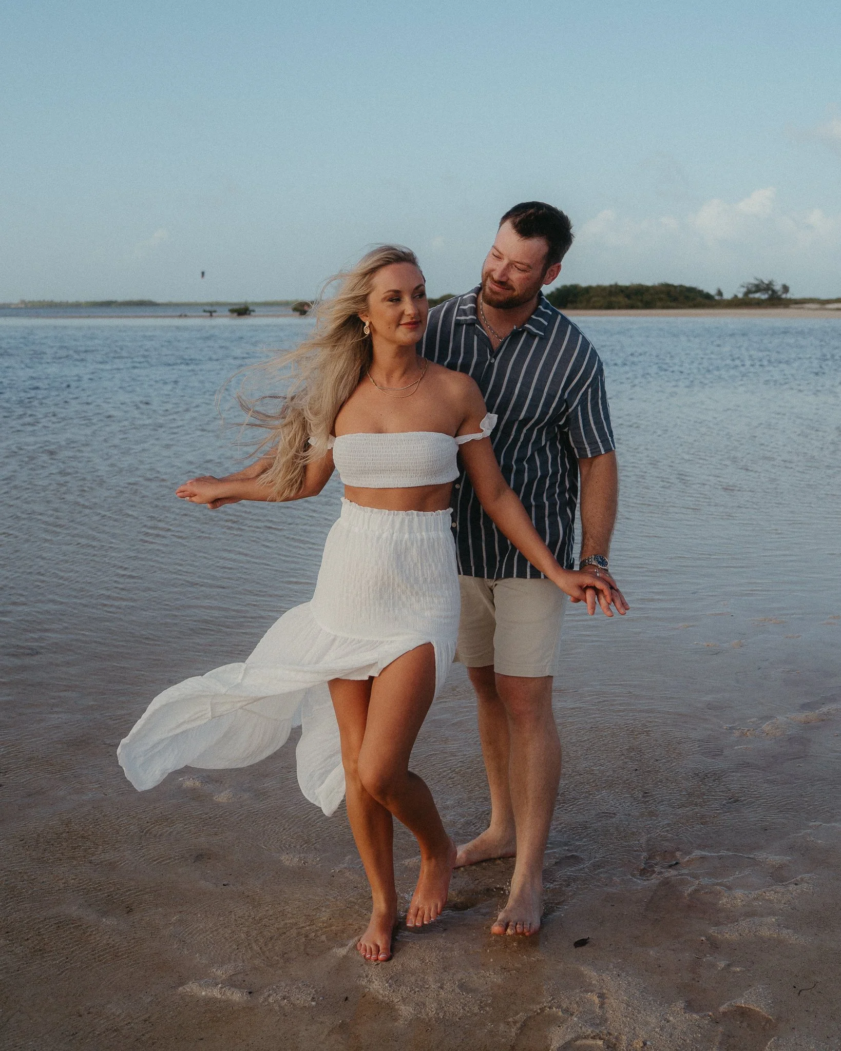 Una pareja caminando por la orilla del mar en un día soleado, disfrutando del paisaje y la compañía, con agua y cielo despejado en el fondo.