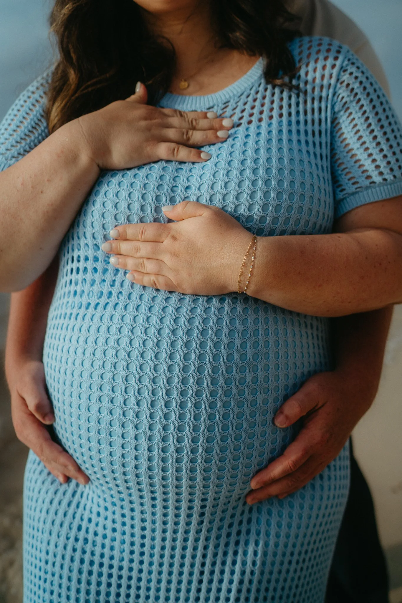 Un embarazo en una mujer con su pareja abrazándola por detrás, ambos colocan las manos sobre su vientre. La mujer viste un vestido azul de malla y lleva una pulsera. La imagen transmite cariño y protección.