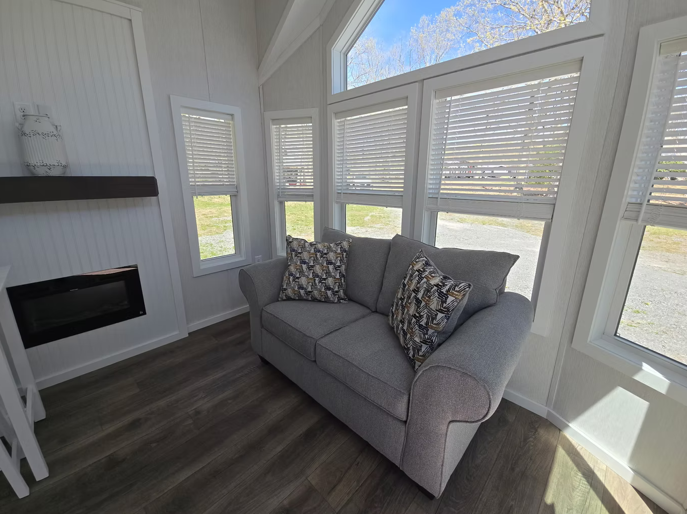 A small living room area with a gray sofa and patterned pillows, surrounded by large windows with white blinds, a dark wood floor, and a white wood-paneled wall with a black shelf and a decorative white vase.