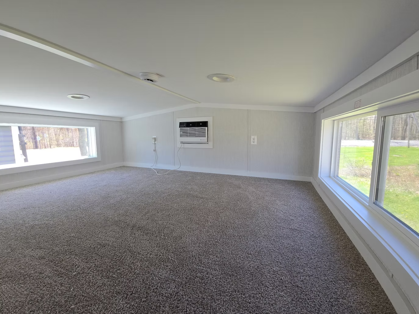 Empty room with large windows, beige carpet, white walls, ceiling air conditioning unit, and window air conditioner.