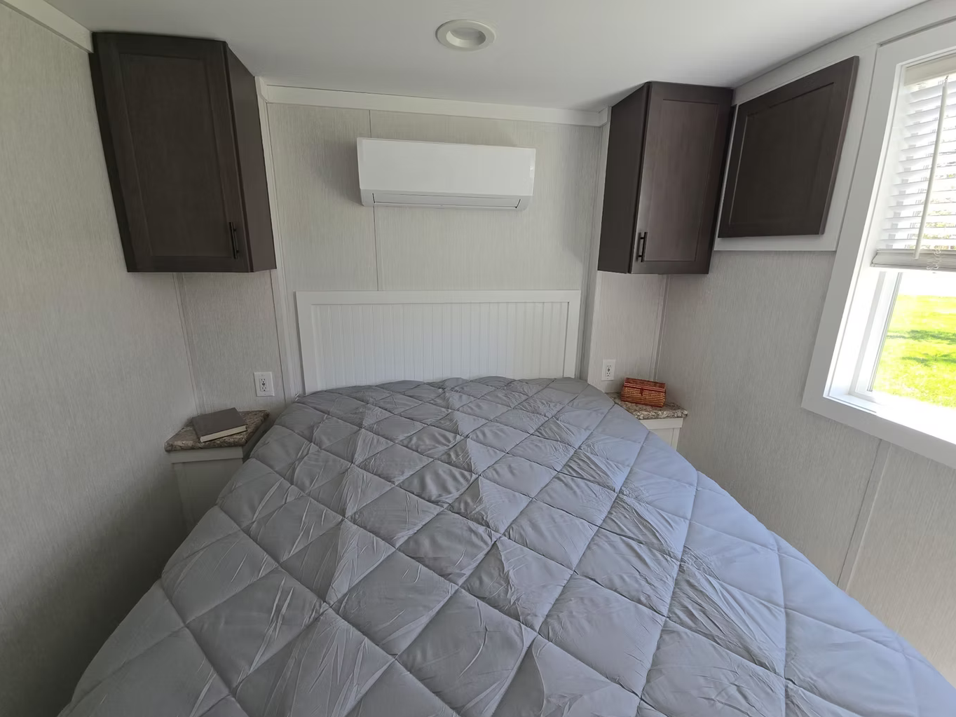 A bedroom with a bed covered in a light gray quilt, two nightstands with items, dark wood cabinets above the bed, a window on the right showing green grass outside, and an air conditioning unit on the wall.
