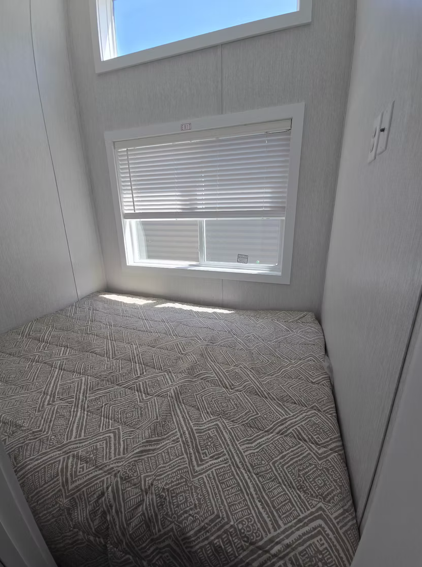 Small bedroom with a window with closed blinds, a bed with a patterned bedspread, light-colored walls, and a corner view.