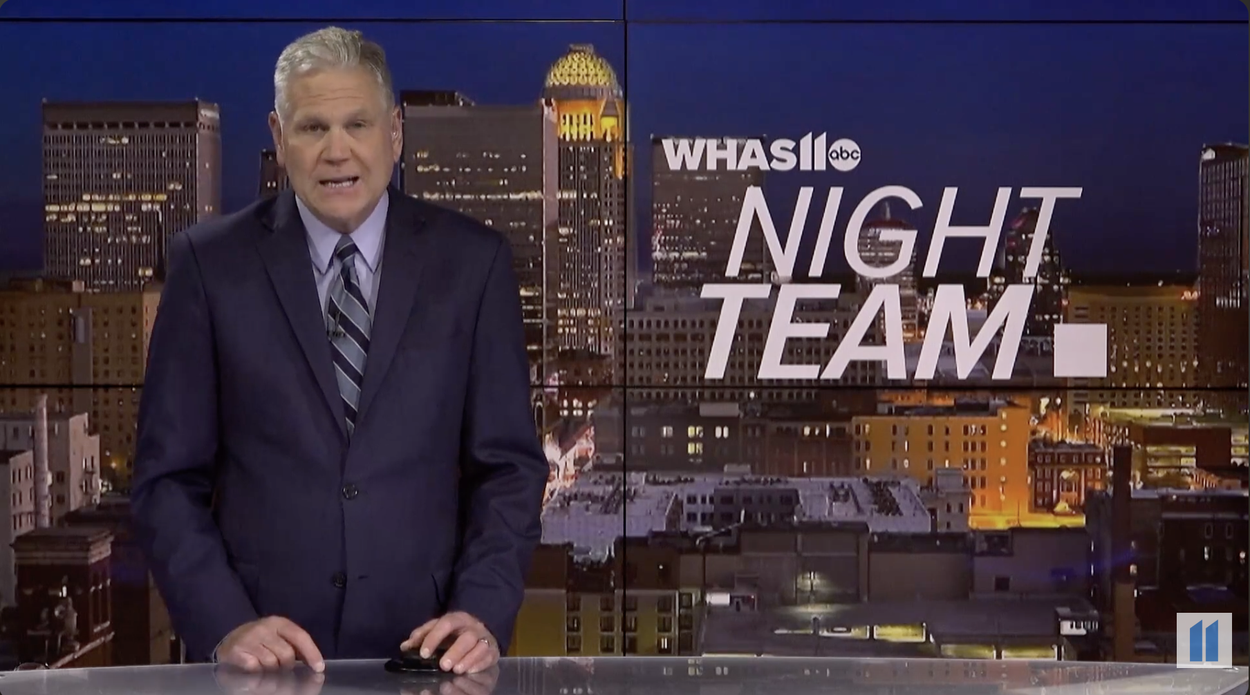 News anchor standing in front of a city skyline at night with a monitor displaying the text 'WHAS11 ABC NIGHT TEAM'.