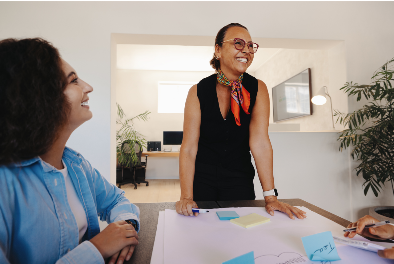 Two women in an office, one standing and smiling, the other sitting and also smiling, with notes and paper on the table.