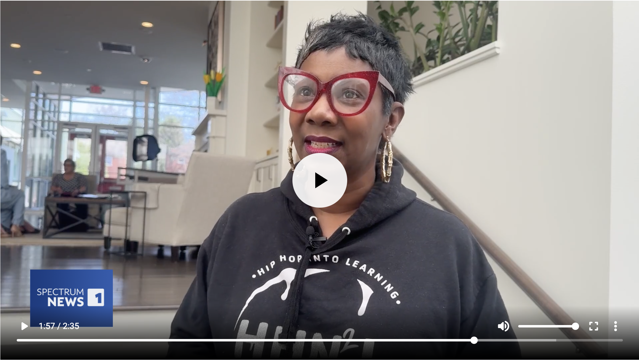 Woman with short black and gray hair, wearing large red glasses and a black hoodie, speaking in a lobby or waiting area with large windows and white walls.