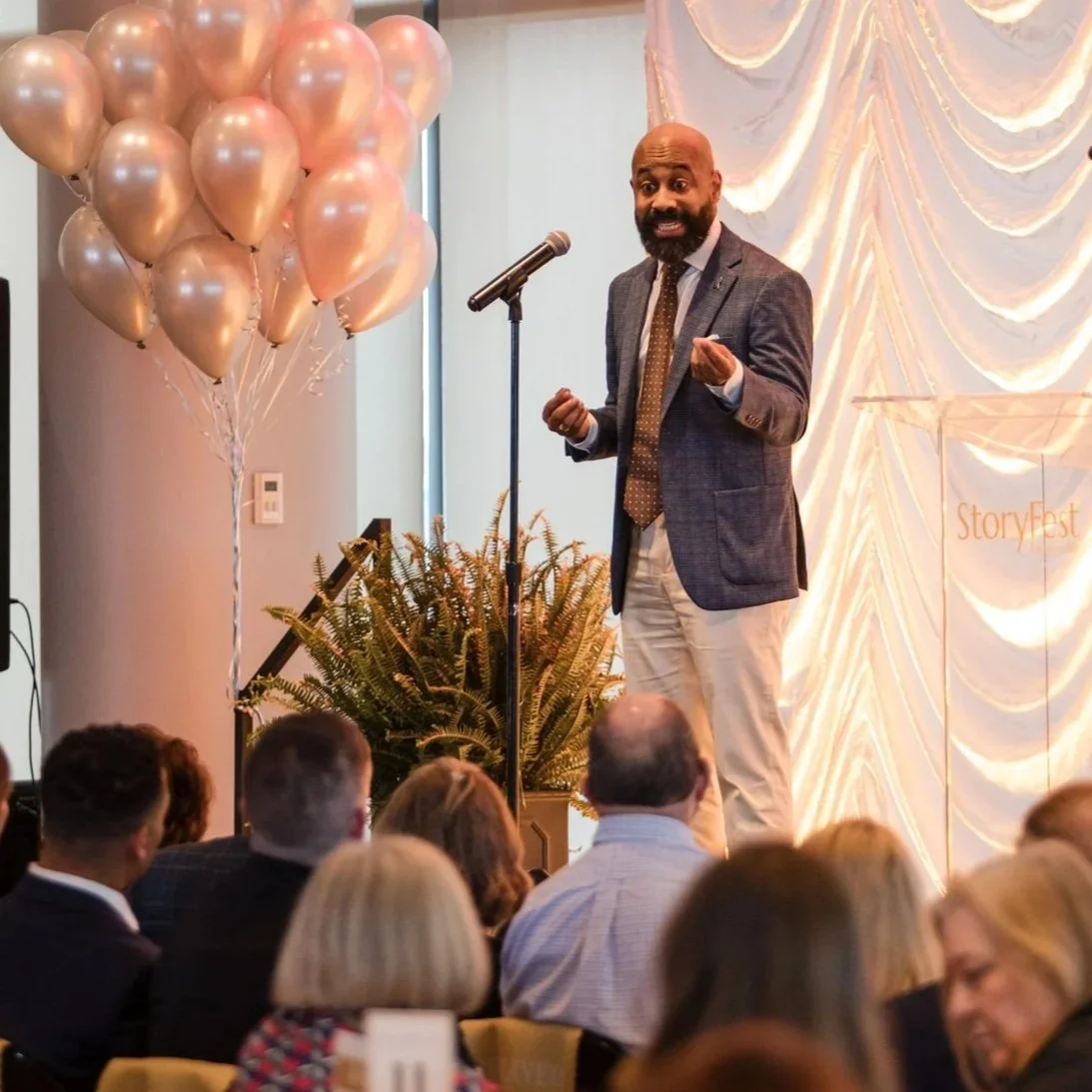 A man in a suit and tie speaking at a microphone during a formal event, with pink balloons and decorative curtains in the background, and an audience listening.
