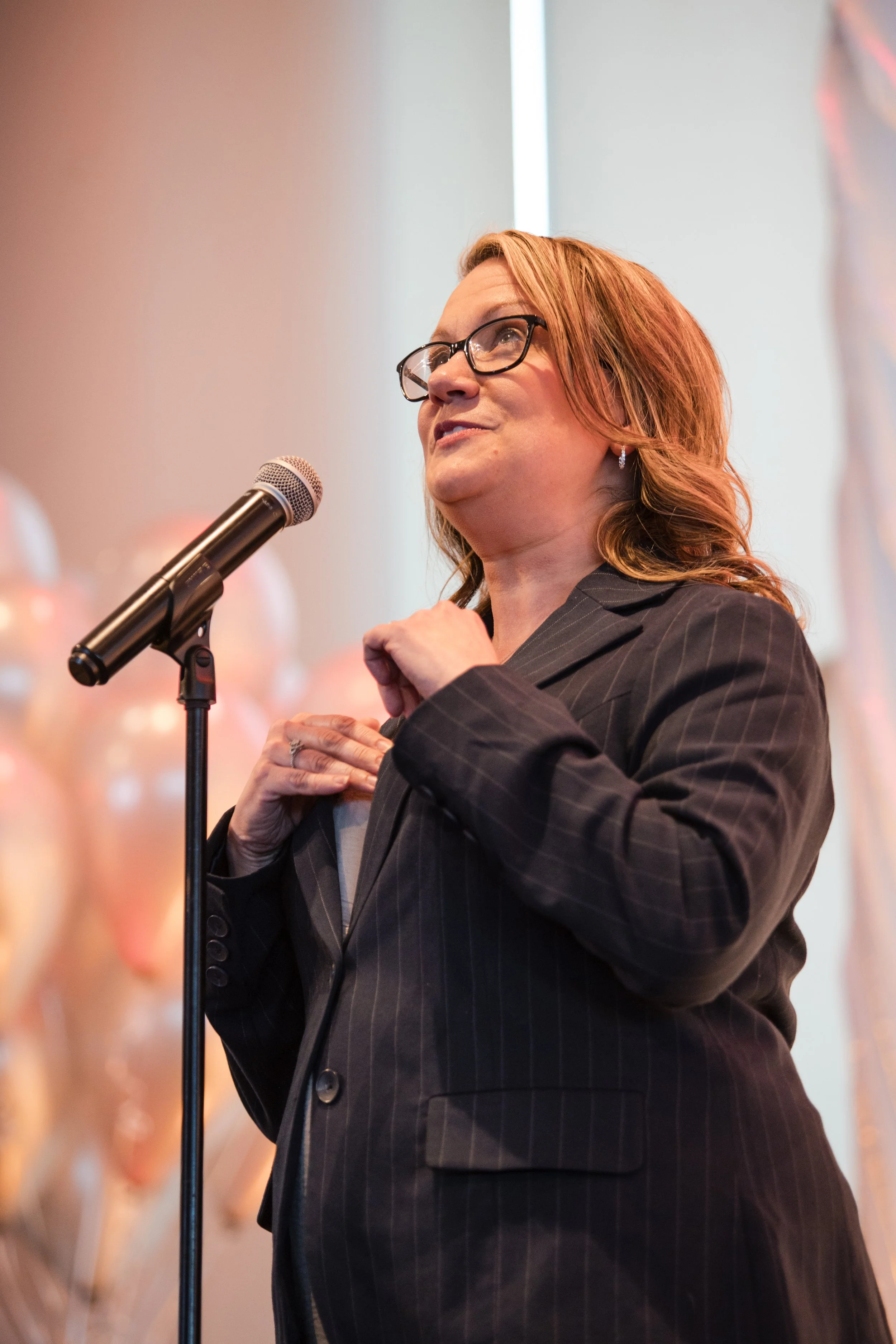 A woman with reddish hair, glasses, and earrings standing at a microphone, speaking or presenting.