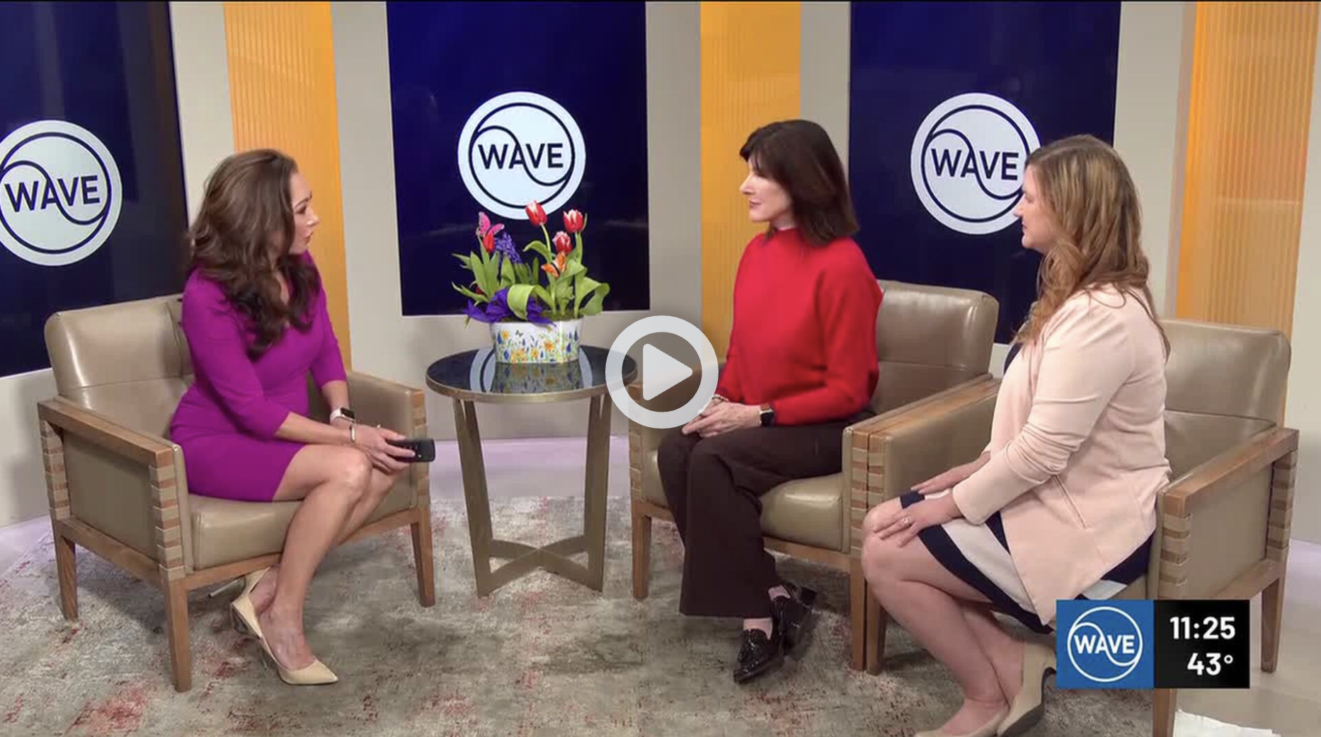 Three women seated on beige chairs having a conversation in a television studio. There is a small round table with a colorful flower arrangement between them and dark blue WAVE logos on the wall behind them. The time displayed is 11:25, and the temperature is 43°F.