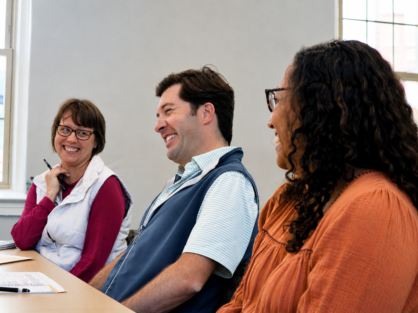 Three people sitting at a table in a meeting room, smiling and engaging in conversation, with papers and a pen on the table.