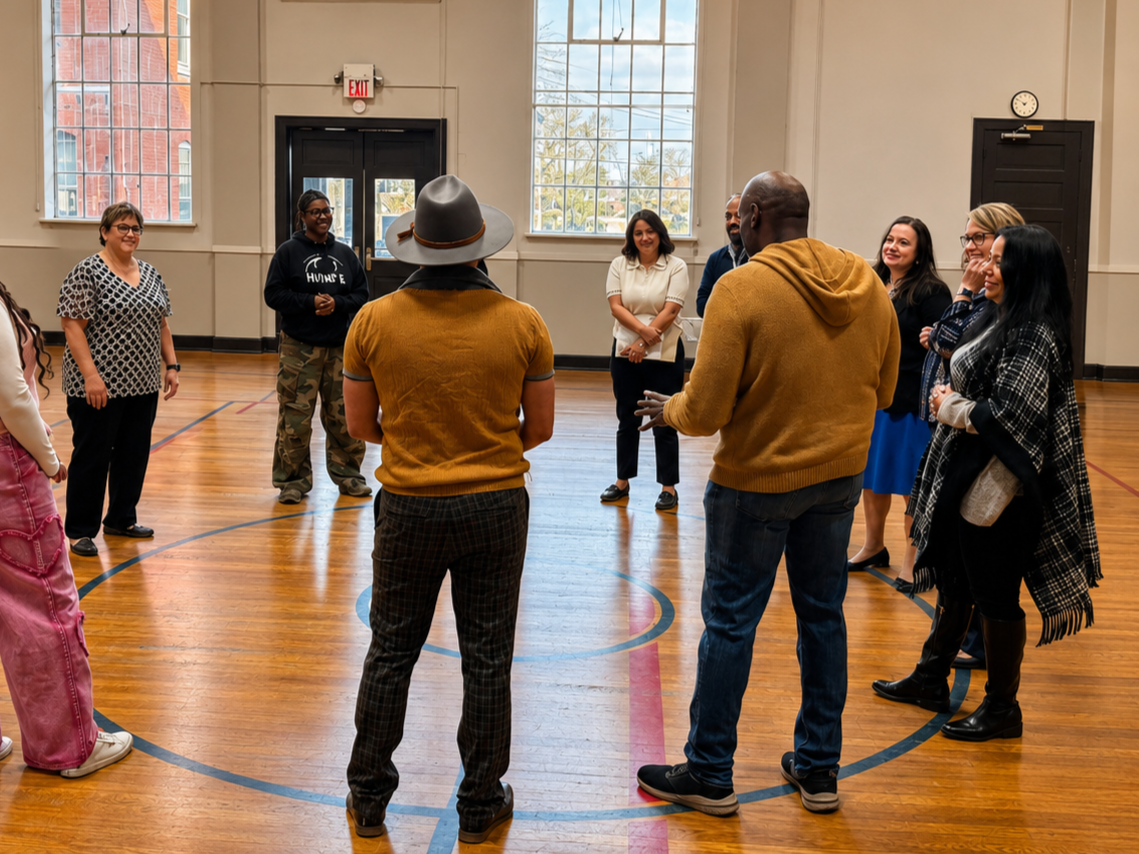 A group of people standing in a circle in a gymnasium, engaging in a conversation or activity. The gym has wooden floors and large windows letting in natural light.