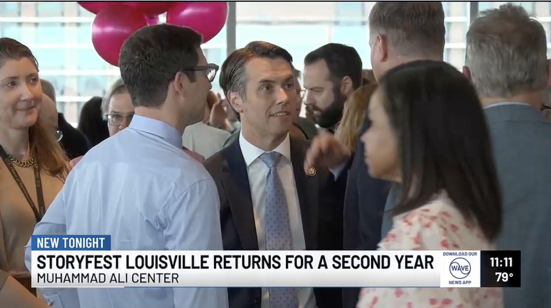 A group of people, including a man in a suit and a man in glasses, are engaged in conversation at an indoor event. Pink balloons are visible in the background, and a news banner at the bottom reads "StoryFest Louisville Returns for a Second Year" at the Muhammad Ali Center, with a time of 11:11 and temperature of 79 degrees.