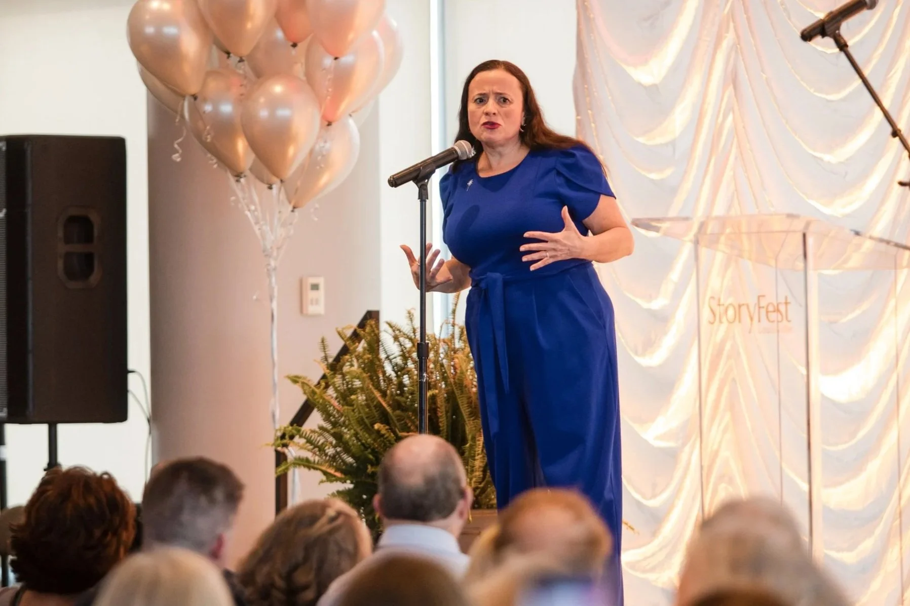 A woman in a royal blue dress speaking into a microphone at an event, surrounded by balloons and audience members.