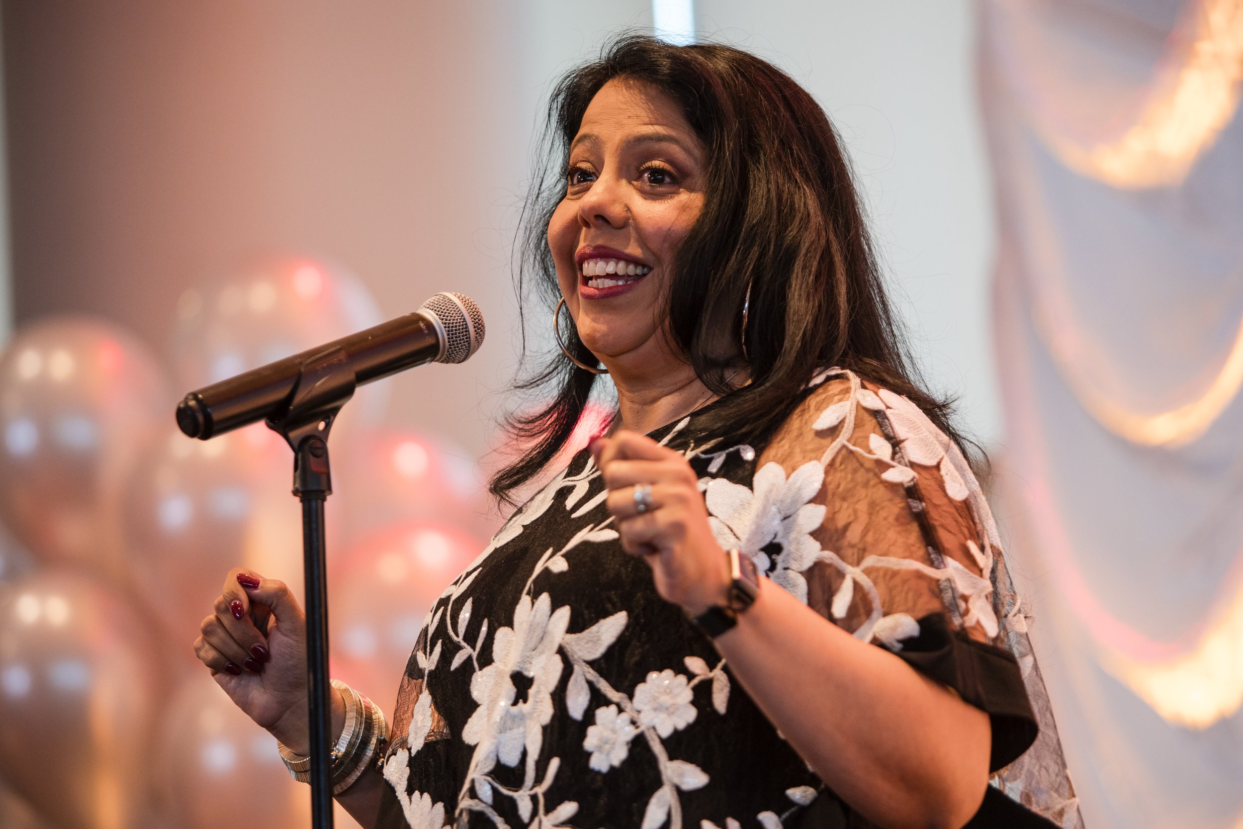 A woman with dark hair and hoop earrings speaking or singing into a microphone at an event with soft lighting and blurred background.