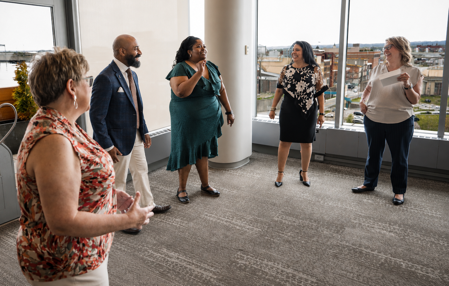 Five diverse adults standing in a circle, engaged in a conversation in a bright office with large windows overlooking a cityscape.