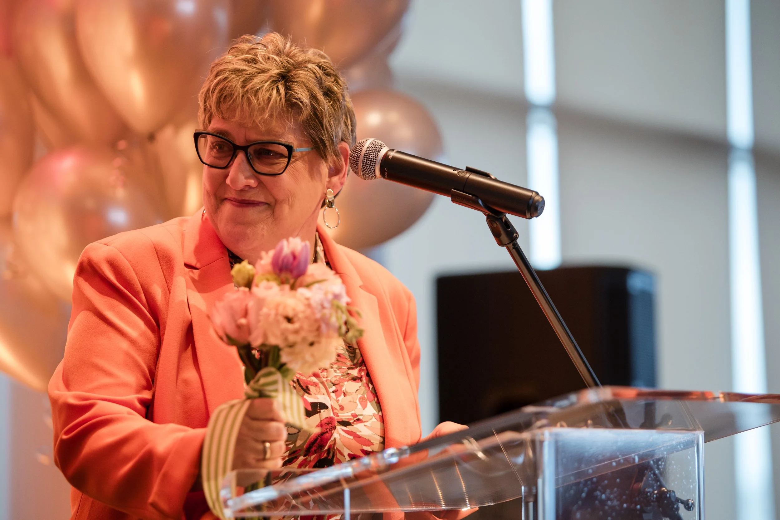 A woman with short, light brown hair and glasses, wearing a peach-colored blazer and floral blouse, holding a bouquet of flowers and standing behind a clear acrylic podium with a microphone in front of her.