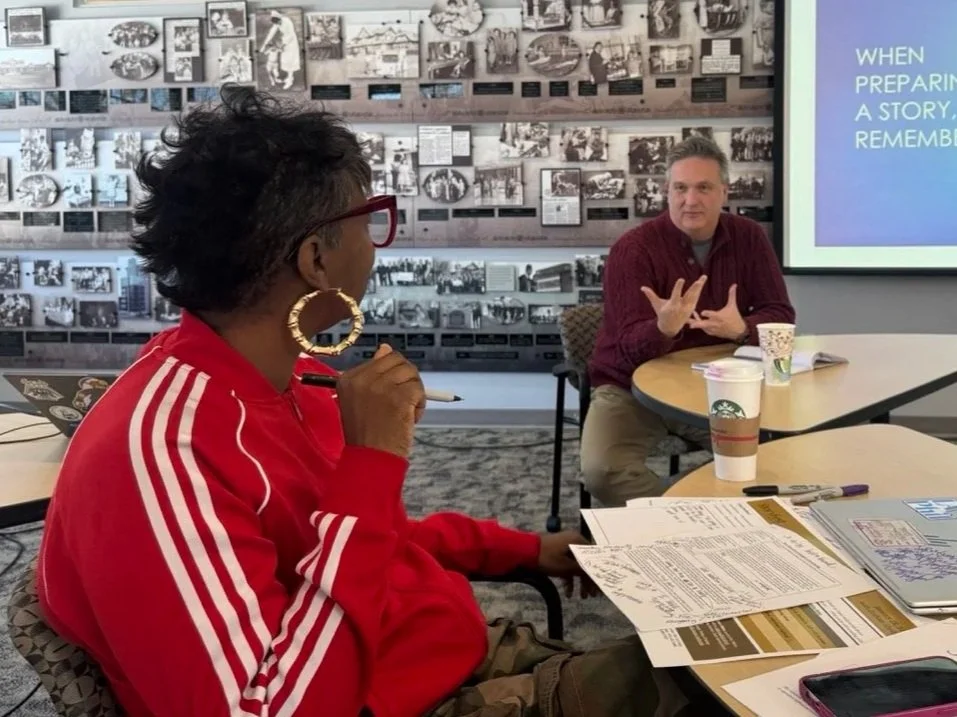 Person in a red jacket with glasses and large hoop earrings listening to a speaker during a meeting in a conference room with photos on the wall and a presentation on a screen.