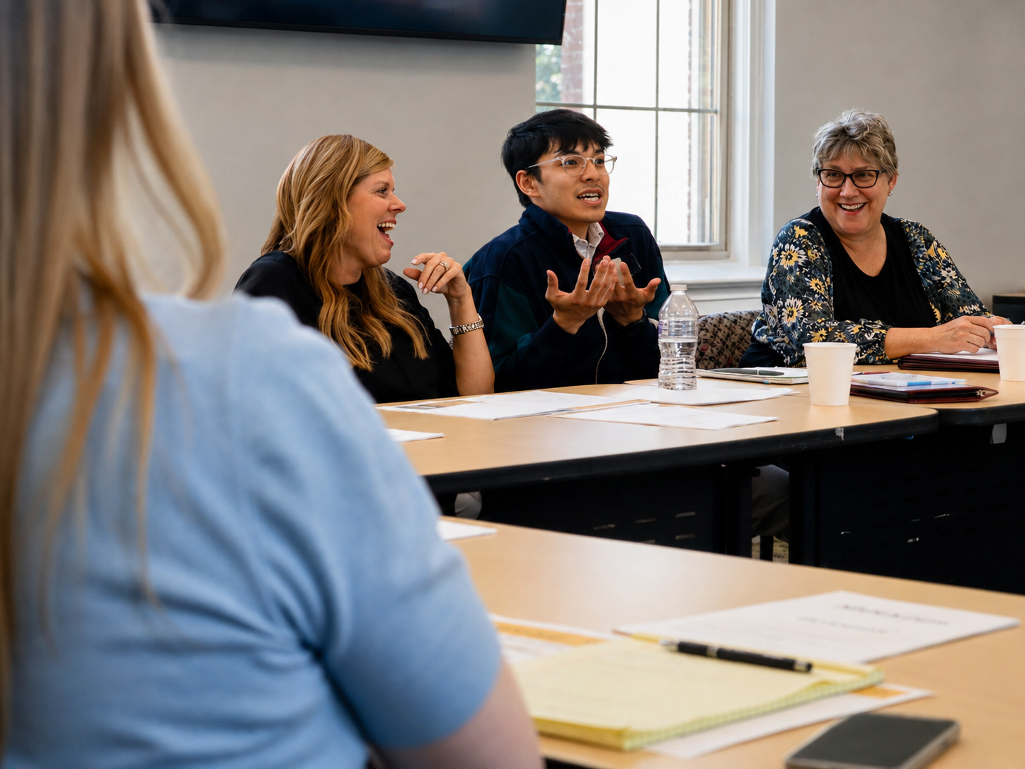 Group of women and a man engaged in a lively discussion at a meeting, with papers, notebooks, and cups on the table.