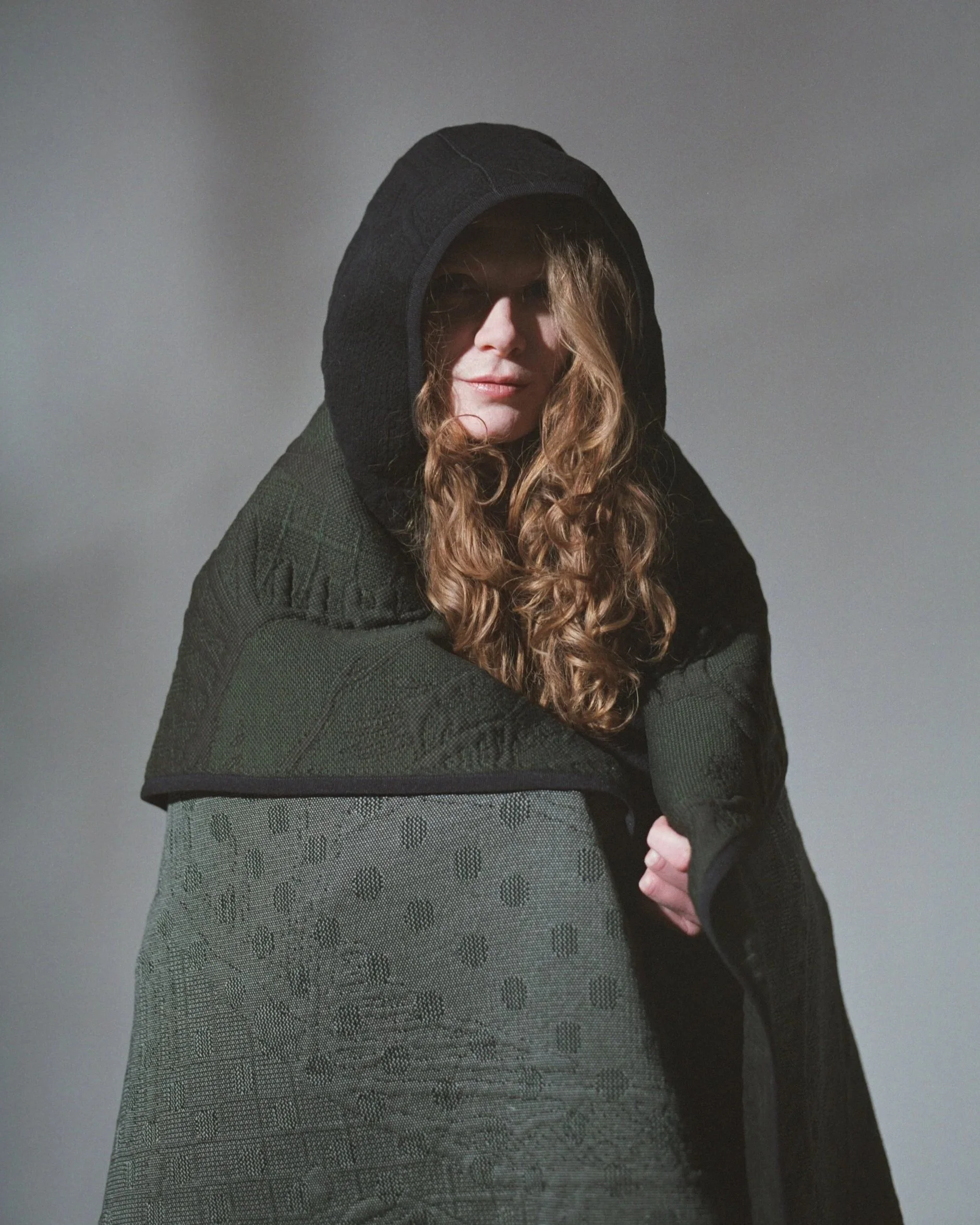 Model wearing a dark green hooded knit, wrapped in a matching 3D knit blanket with embossed tattoo-inspired patterning. Textile development by Byborre in collaboration with Henk Schiffmacher.