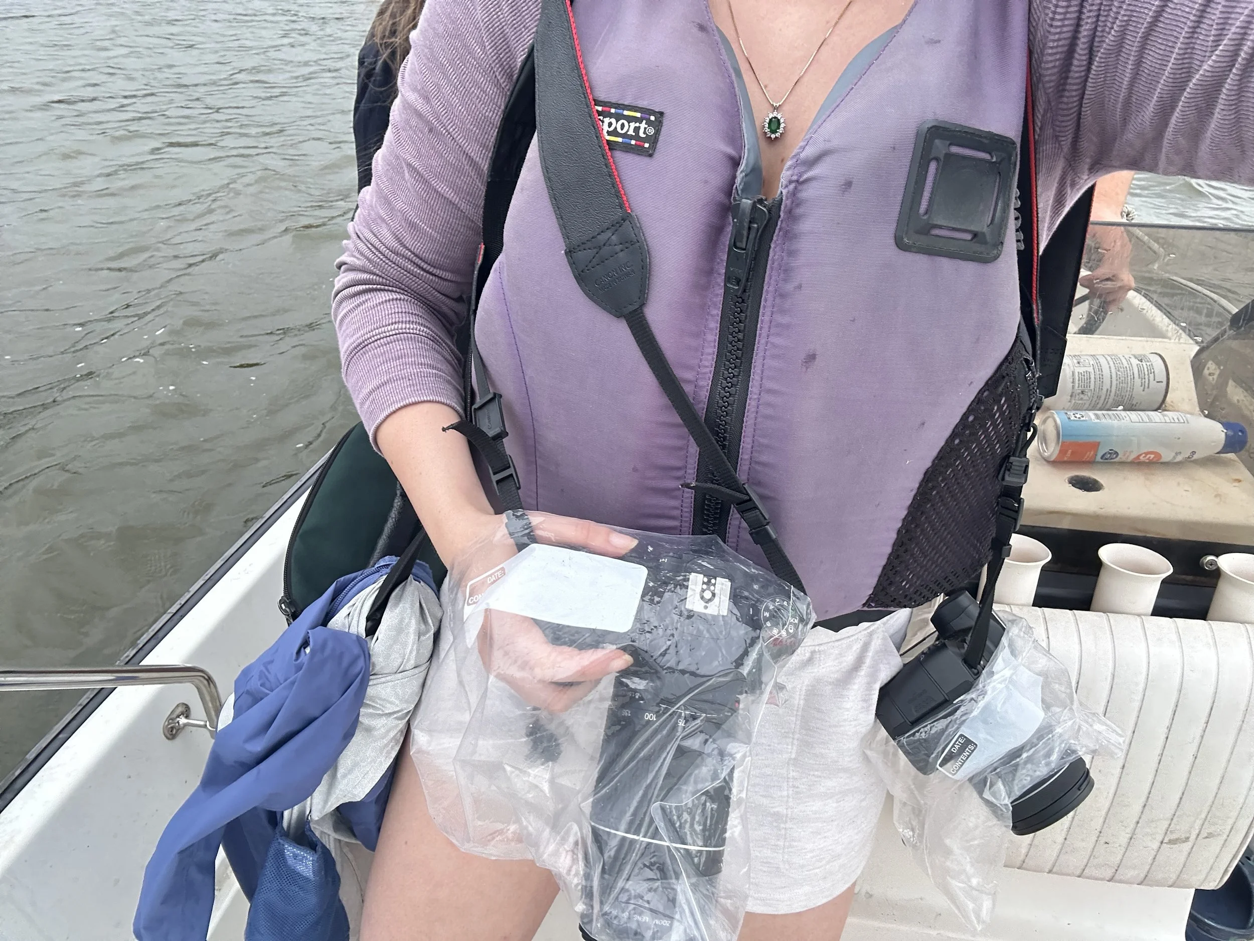 Person on a boat holding a plastic bag with a label, wearing a purple backpack and a purple jacket, with water in the background and spray cans on the boat deck.