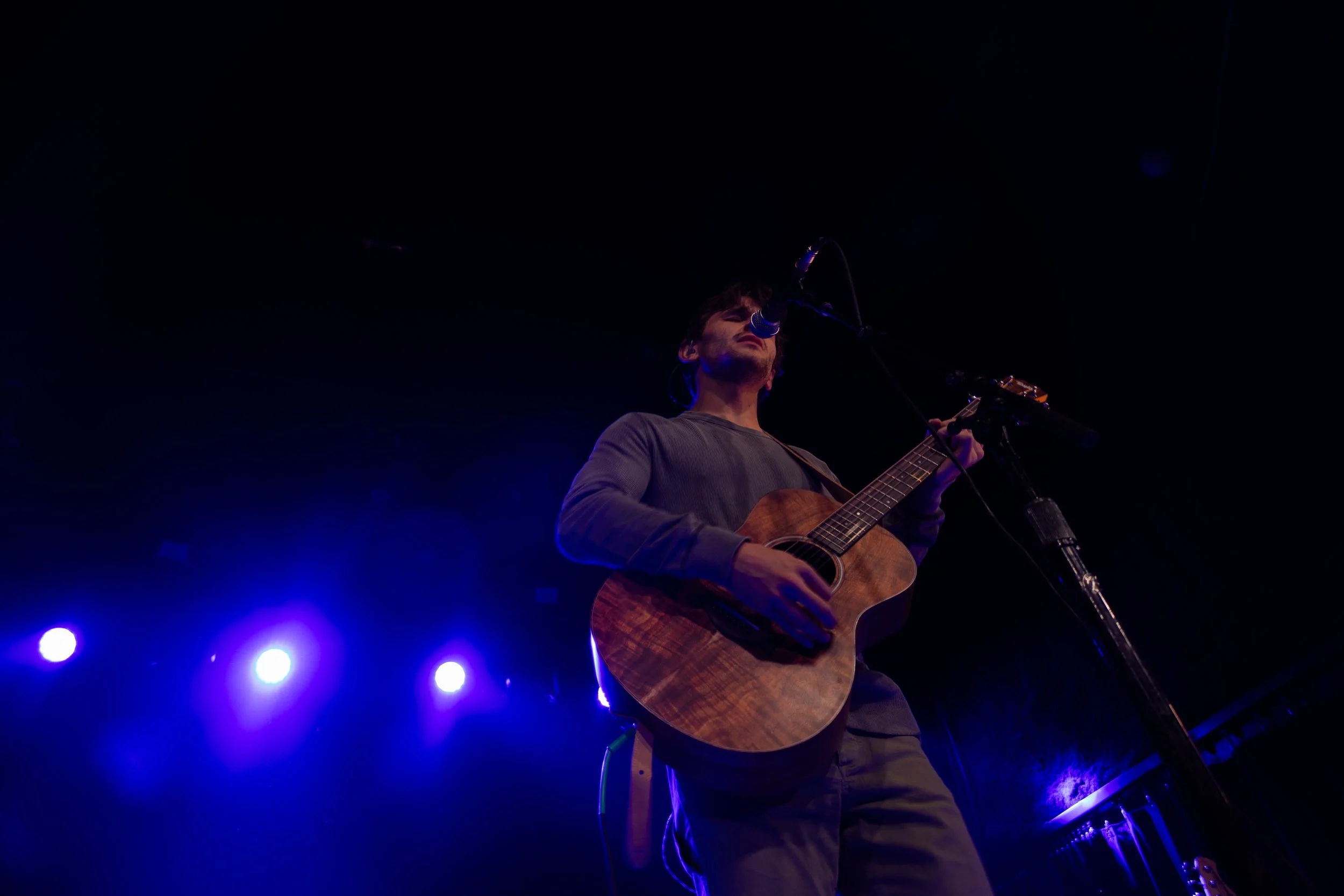 A man playing an acoustic guitar and singing into a microphone on stage, with blue stage lights shining in the background.