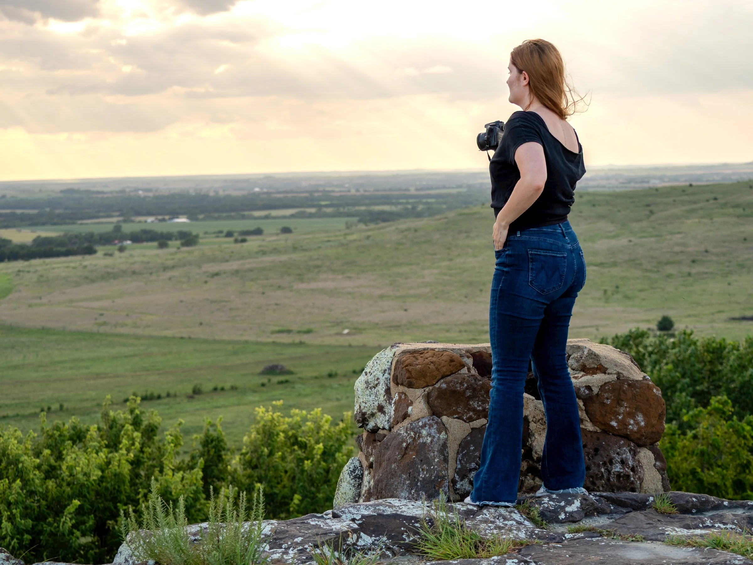 A woman with red hair wearing a black shirt and blue jeans stands on a stone wall, looking out over a green field with a cloudy sky in the distance.