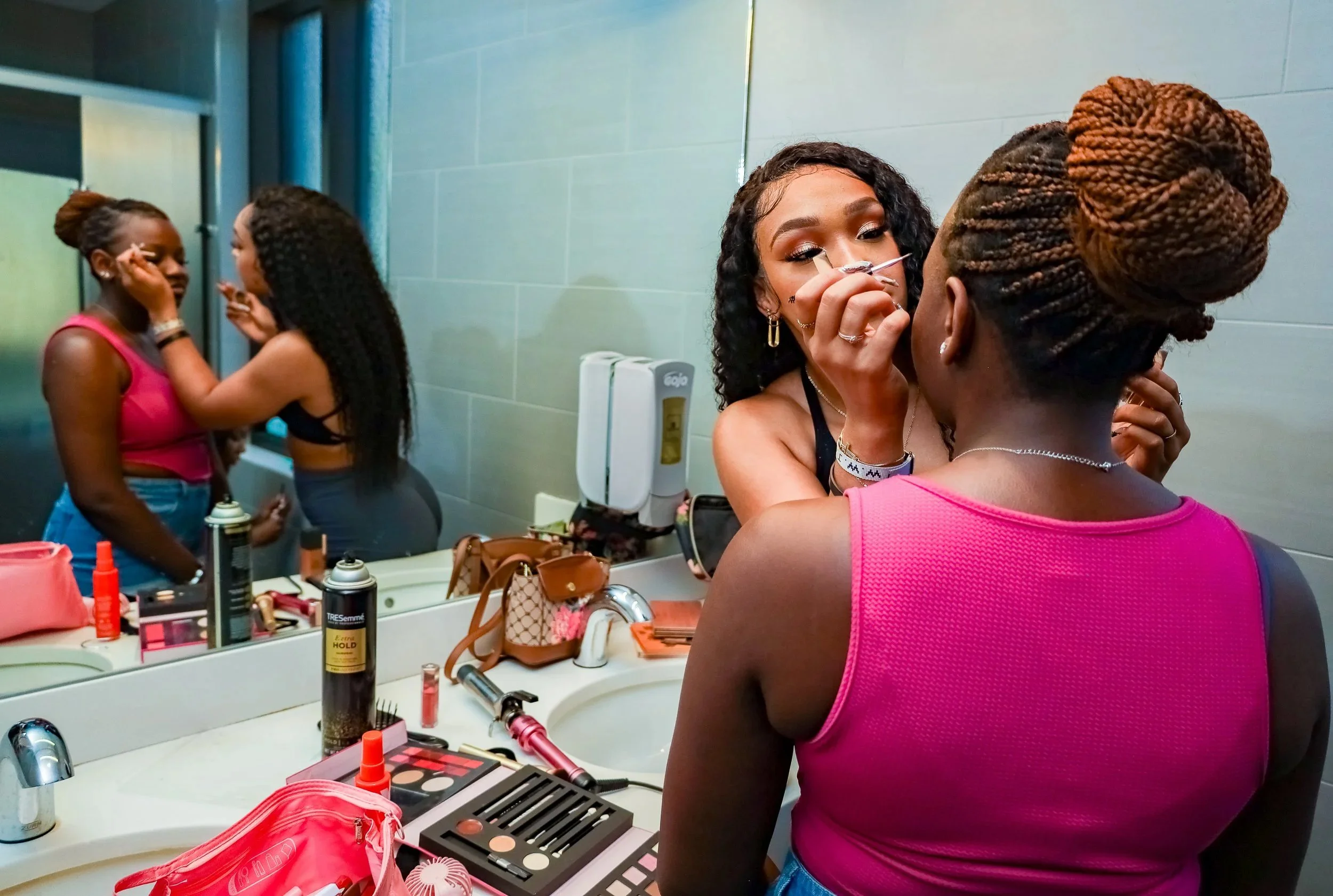 Two women getting makeup done in a bathroom, with various makeup products on the counter and a mirror reflecting the scene.