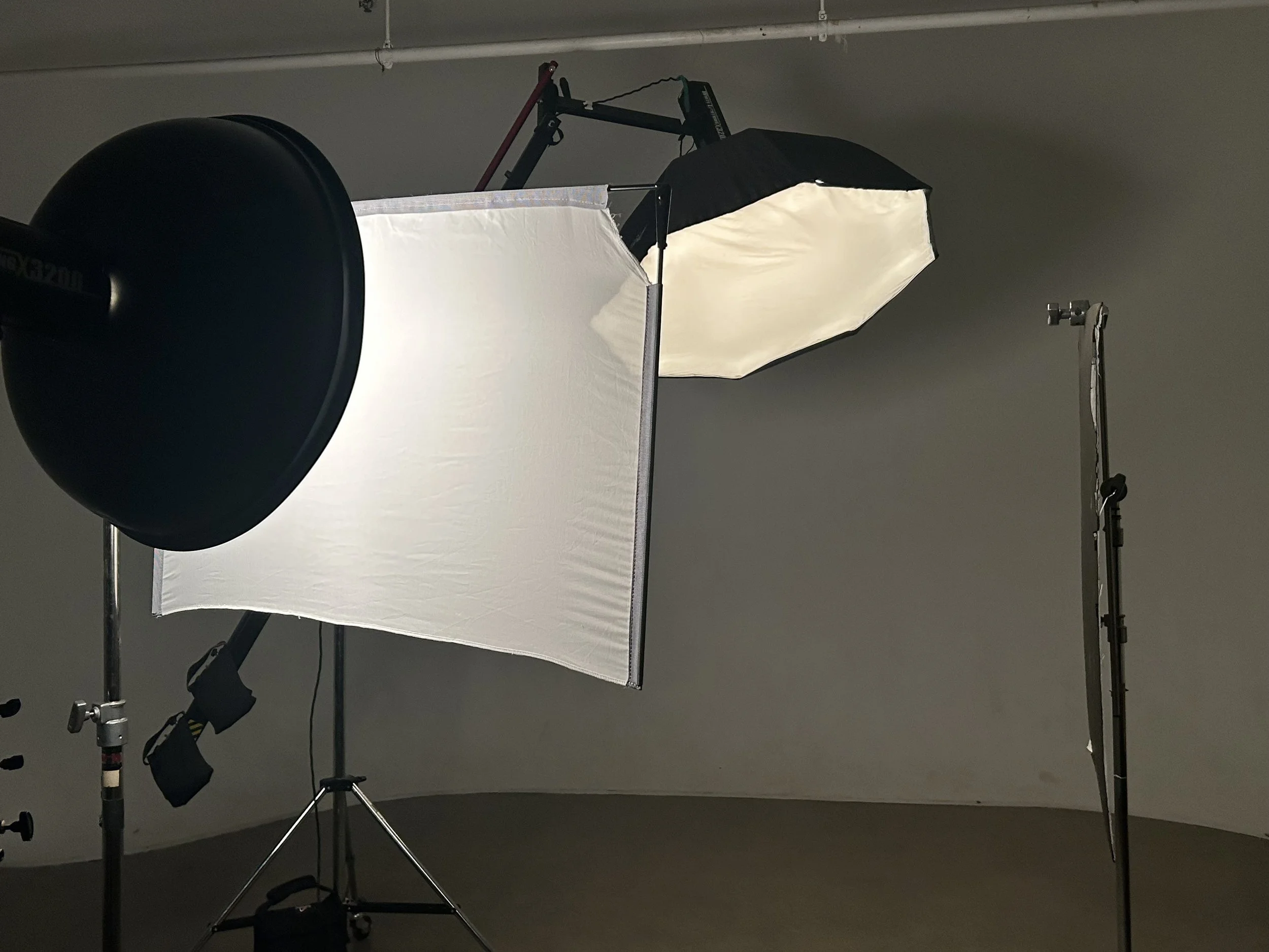 Photography studio setup with softbox lighting, a large reflector, and studio stands against a plain wall.