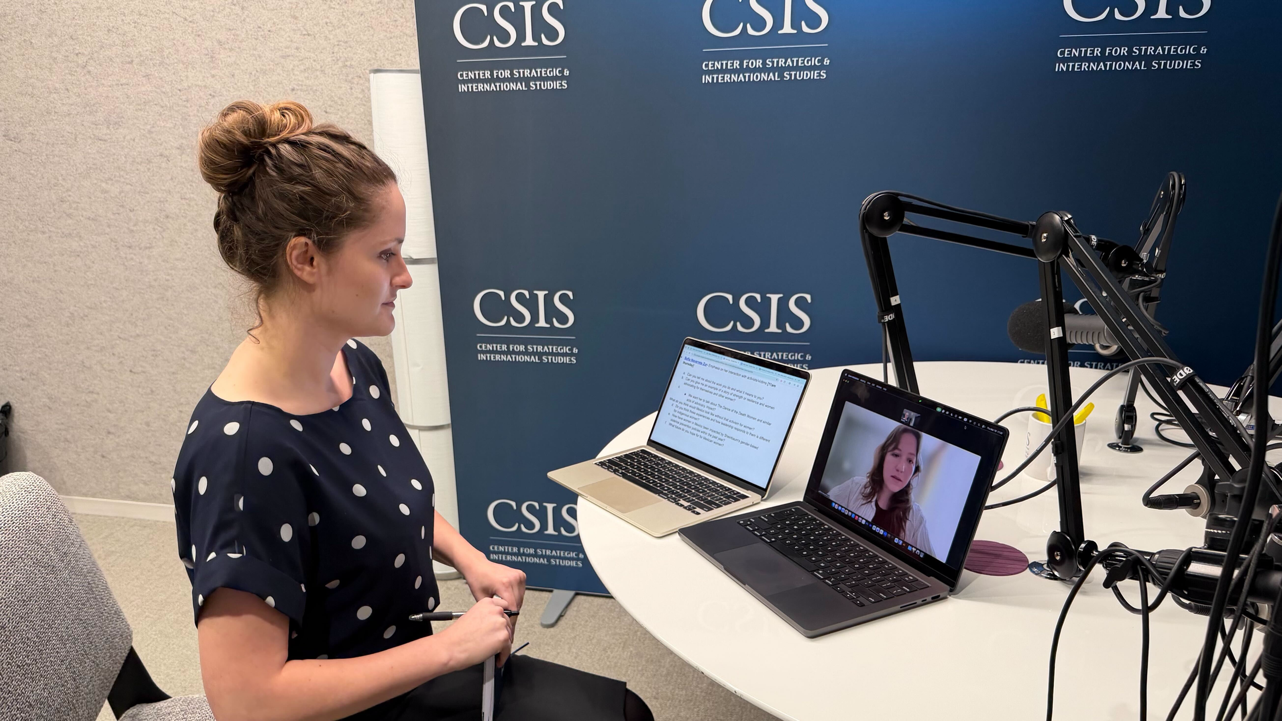 A woman with a bun hairstyle and polka dot blouse sitting at a table with laptops, during a virtual meeting at the Center for Strategic & International Studies.