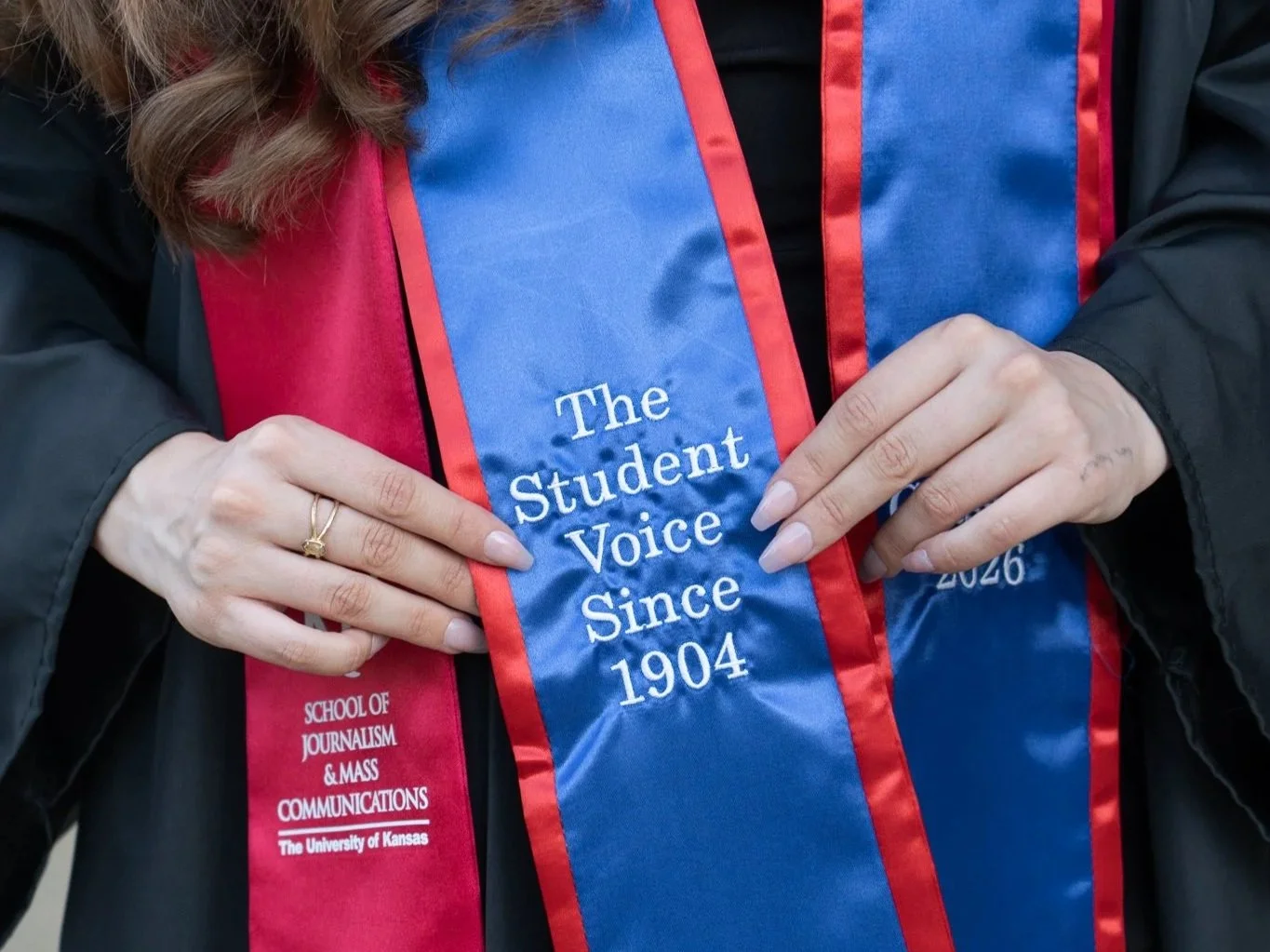 Person holding a sash that reads "The Student Voice Since 1904," wearing a black jacket and a pink and blue academic stole from The University of Kansas School of Journalism & Mass Communications.