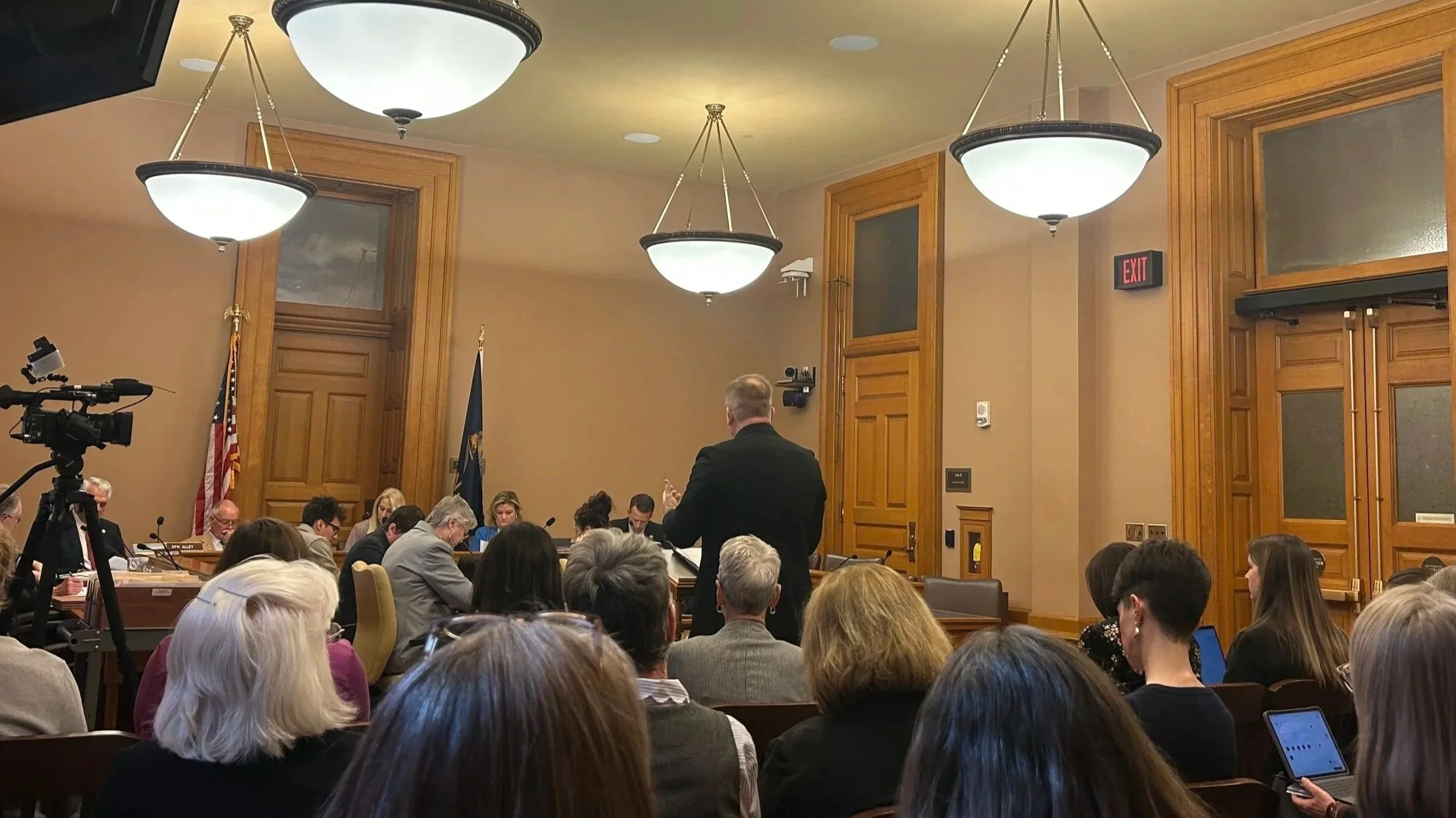 A crowded conference room filled with people sitting at tables, many looking towards a speaker standing at the front. The room has wood-paneled walls, large light fixtures, and an exit sign visible.